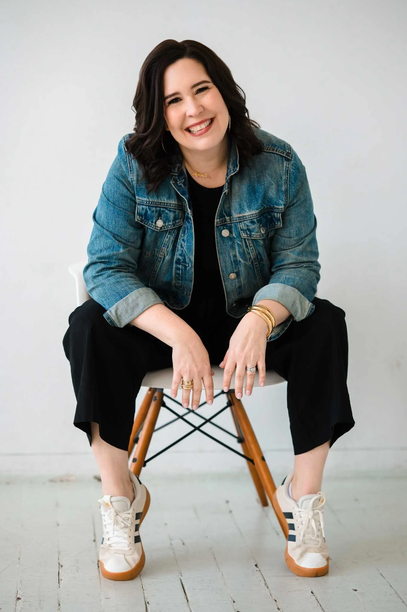 A woman with dark wavy hair smiling and squinting, sitting on a white chair with wooden legs against a plain light-colored background, wearing a denim jacket, black pants, white sneakers, and gold jewelry.