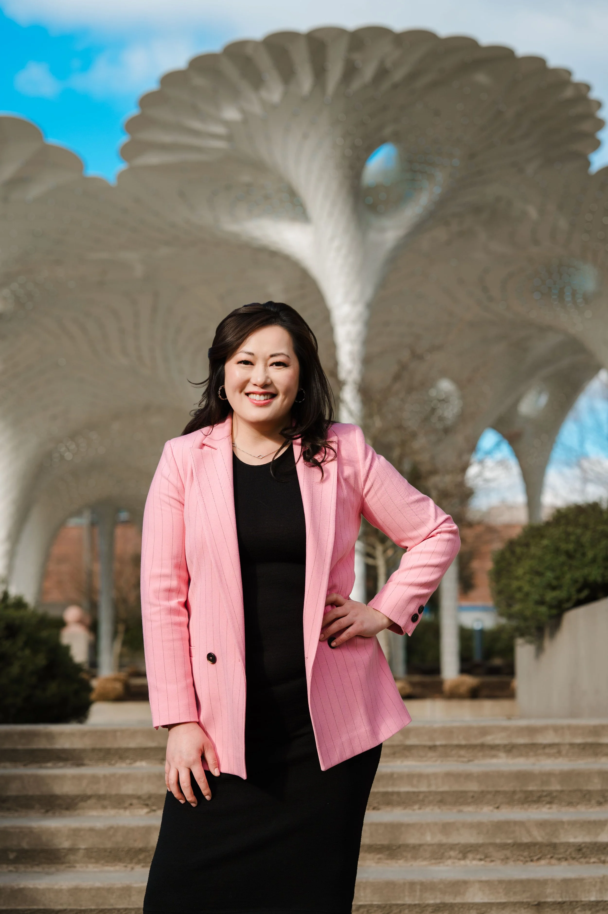 Woman in a pink blazer and black dress smiling in front of a modern, metallic sculpture outdoors.