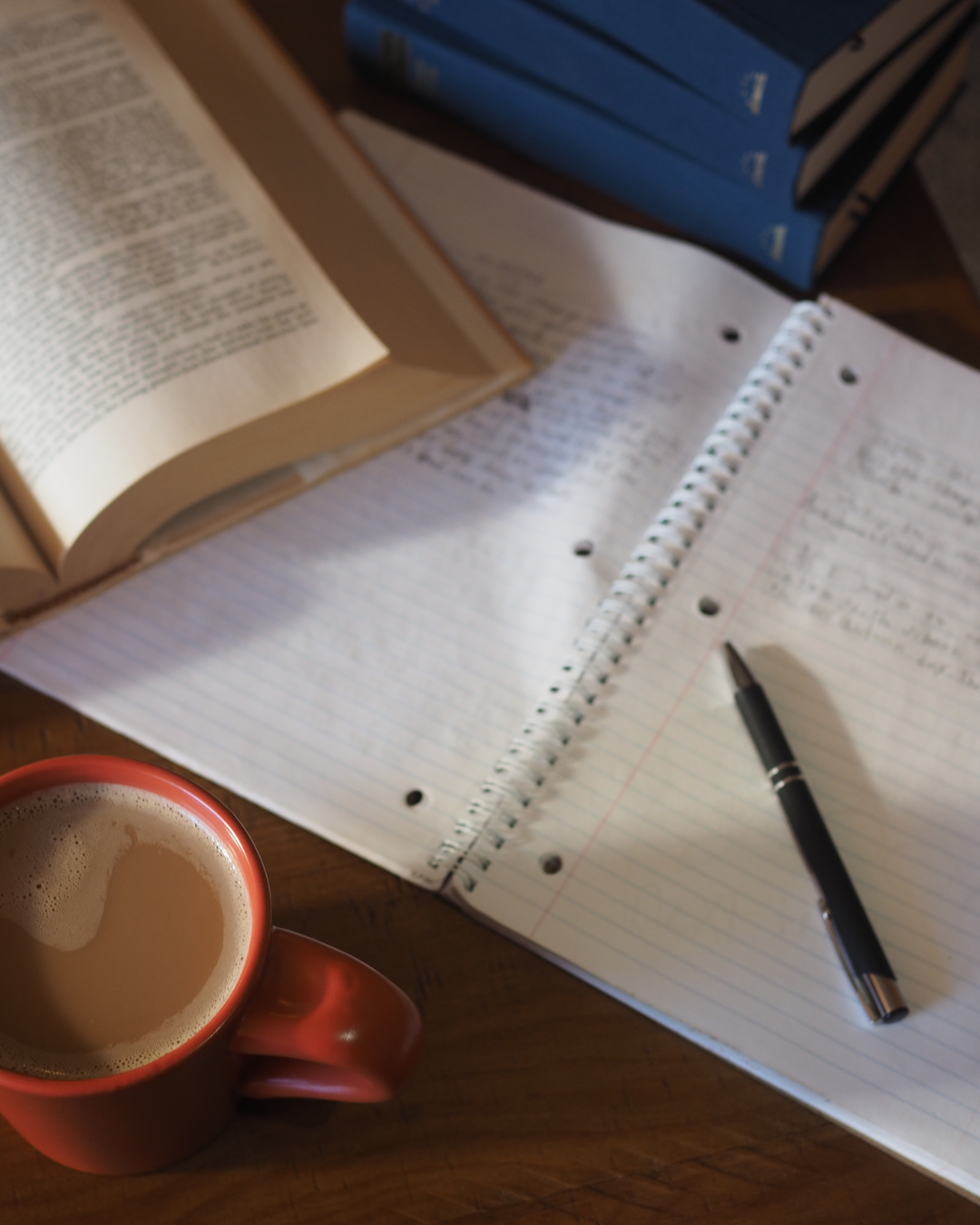 Open notebook with handwritten notes, a black pen, a cup of coffee, and open books on a wooden surface.