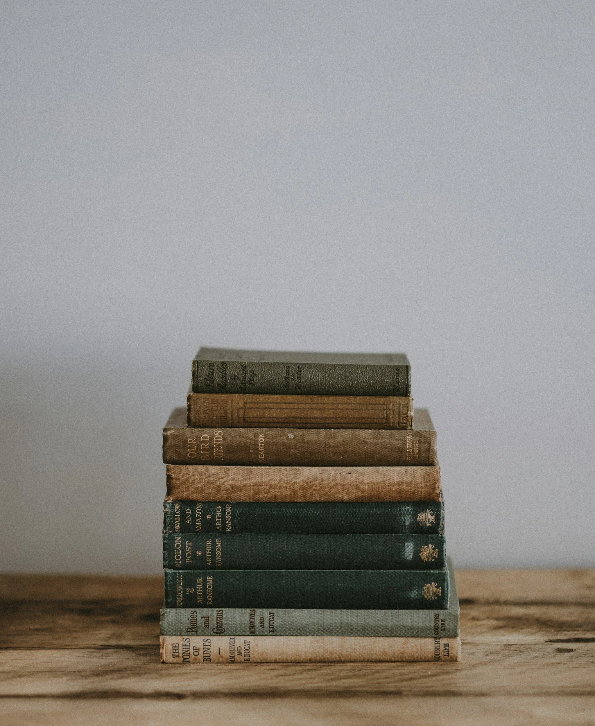 A stack of old hardcover books arranged vertically on a wooden surface, with a plain gray background.