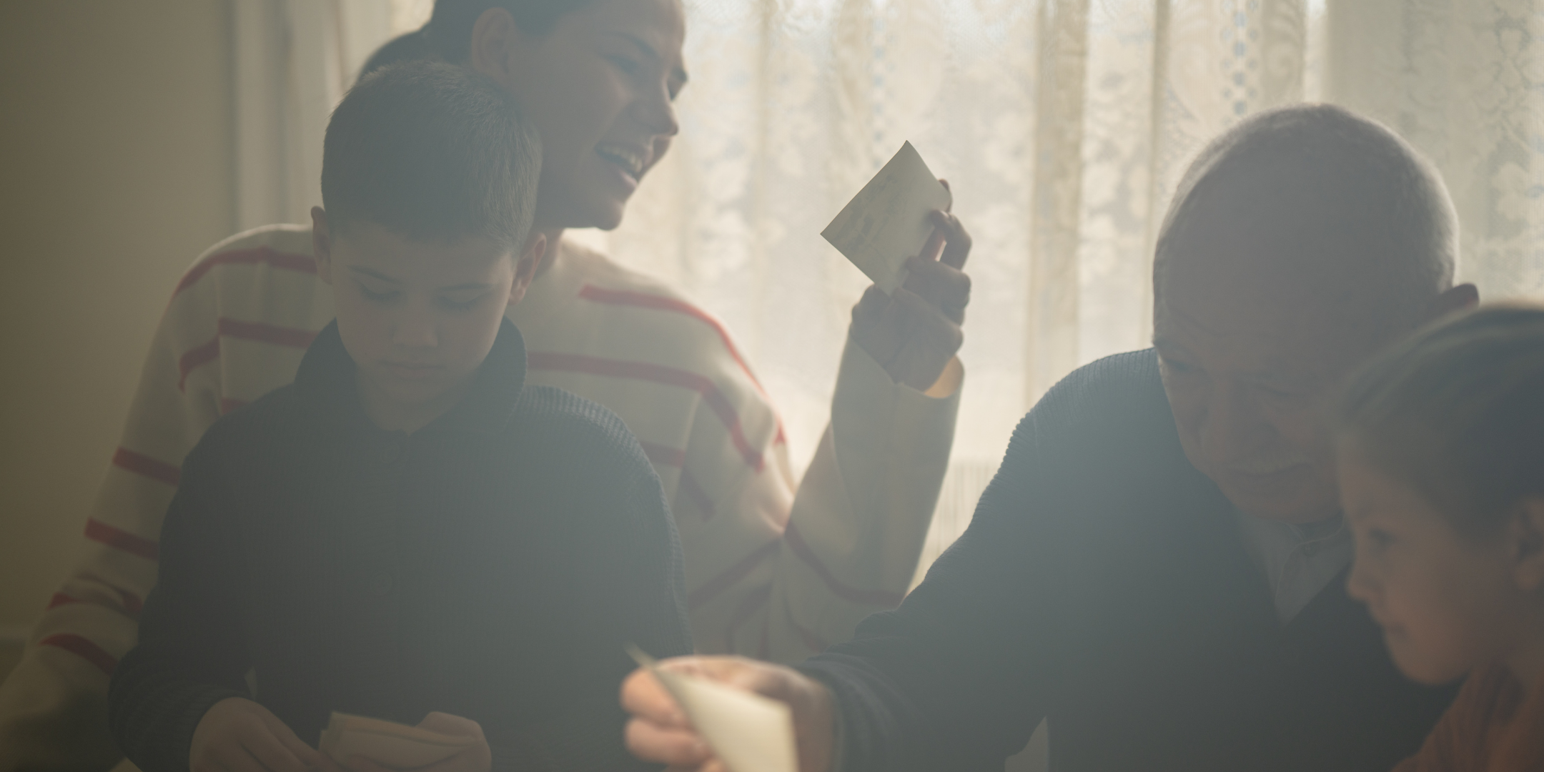 Family gathered indoors, smiling, sharing a moment, with a woman holding a mug, and a child and older woman nearby.