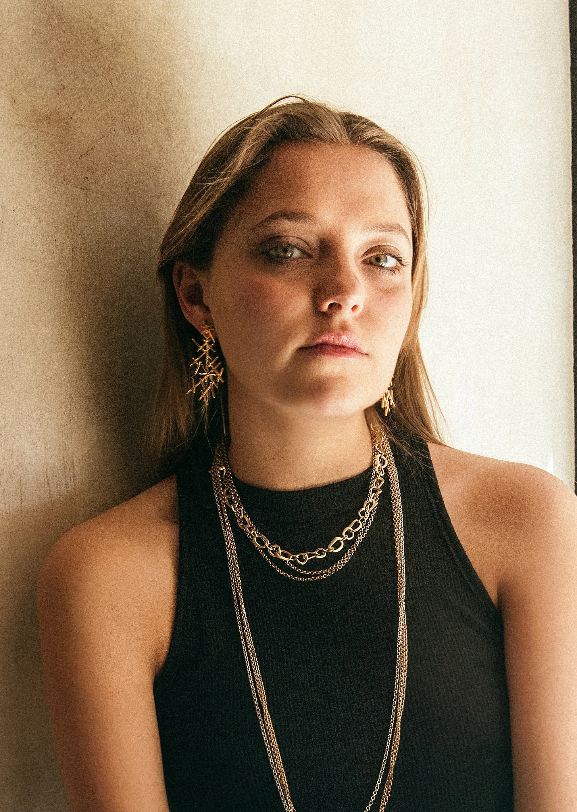 The image depicts a white woman with long dark blonde hair, adorned with various types of gold earrings and necklaces. Showcasing her stylish and sophisticated fashion sense, she is wearing a simple black vest.