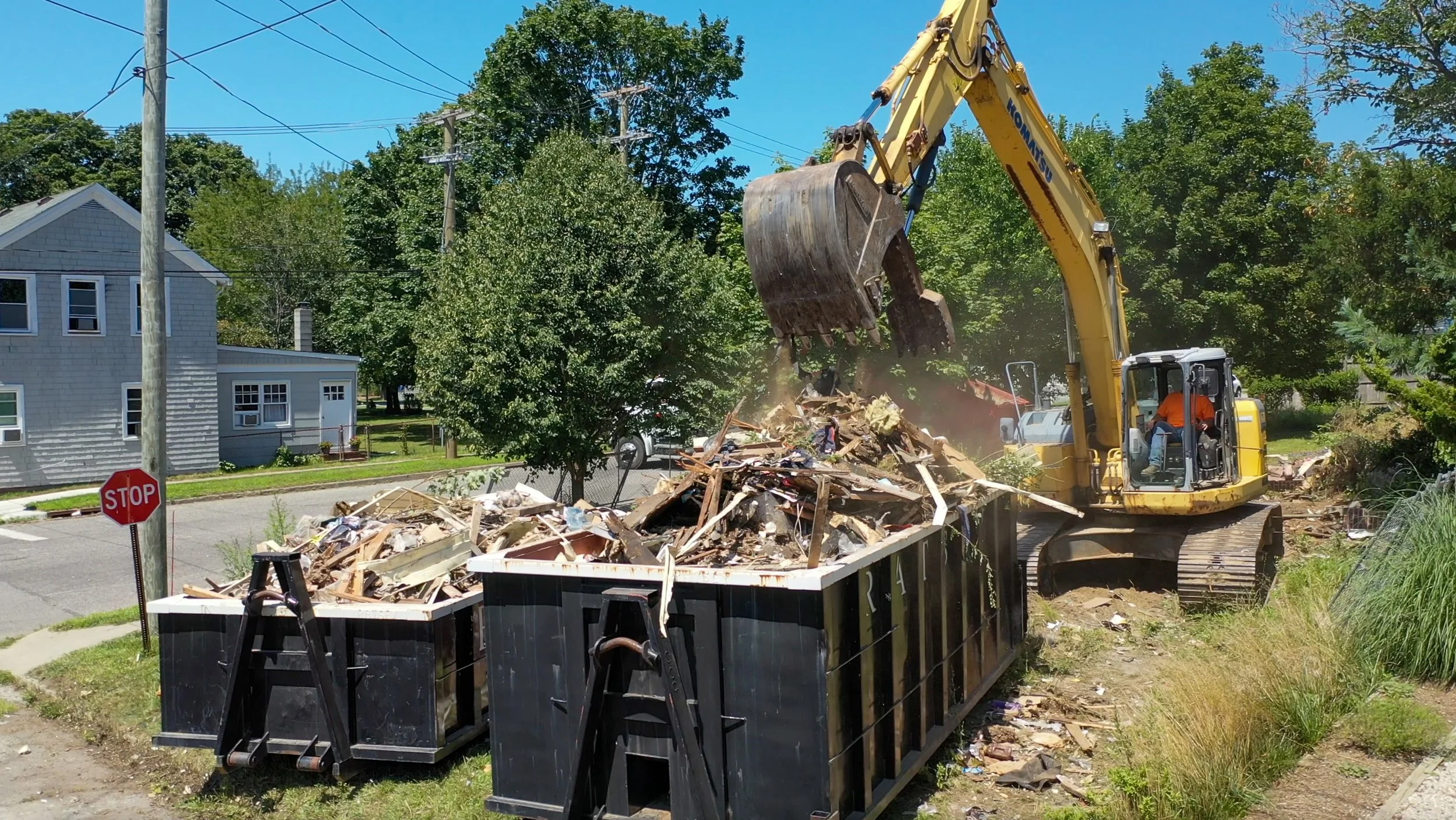 An excavator with a yellow arm and bucket is picking up debris from a large black dumpster filled with construction or demolition waste, on a sunny day near residential houses and trees.