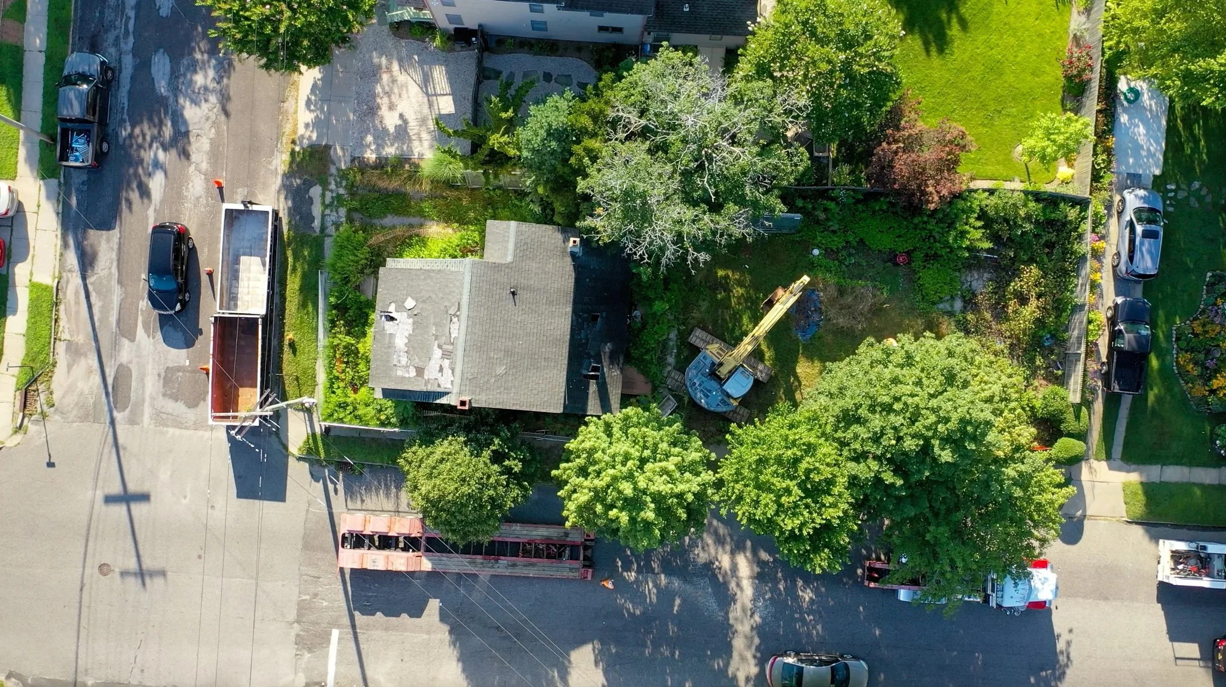 An aerial view of a residential area with a house surrounded by lush green trees, a small yard, parked cars on the street, a construction crane near the house, and a cleared driveway.