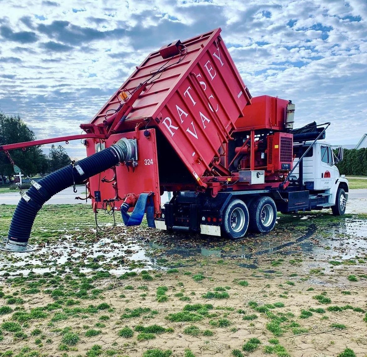 A red street sweeper truck with the words "RATSEY SERVICE" on its side, parked on a grassy area with puddles of water and a partly cloudy sky in the background.