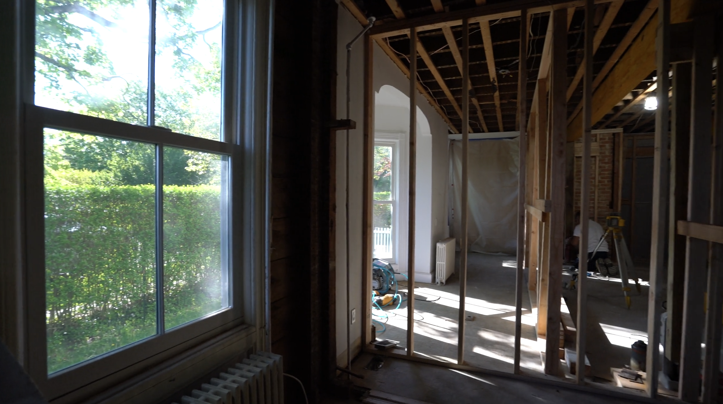 Interior of a house under renovation (by Ratsey Construction) with exposed wooden framing, construction tools, and a large window showing greenery outside.