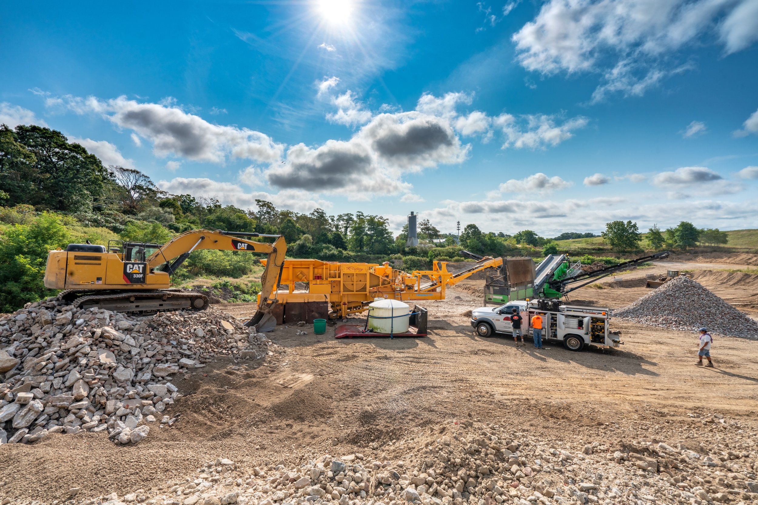 Ratsey Construction site with excavator, rock crusher, gravel, and construction workers under a partly cloudy sky.