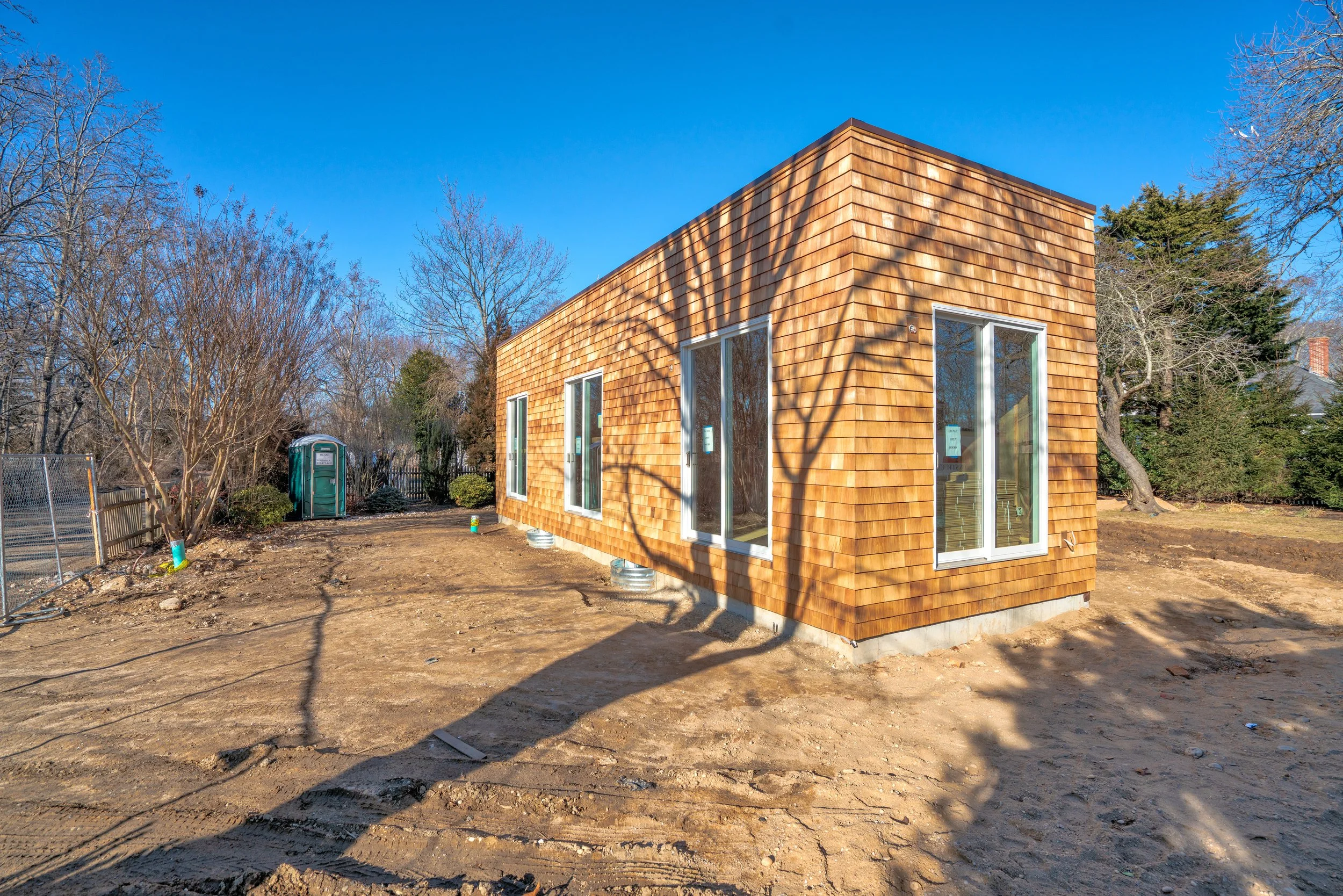 A modern small wooden house with large glass doors and windows on a construction site, surrounded by bare trees and dirt ground.