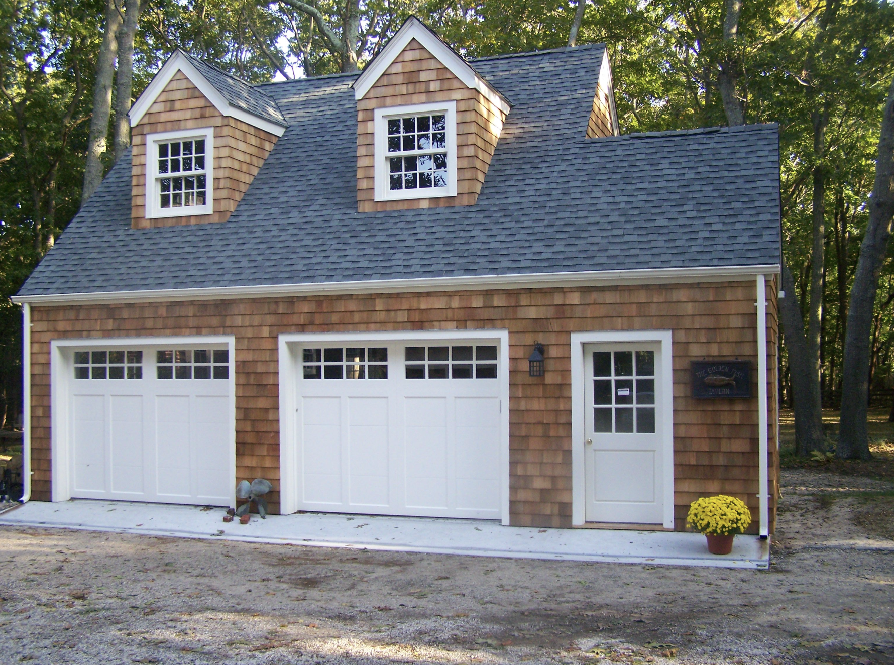A two-story garage with a blue shingled roof, wooden shingles on the upper section, white garage doors, a white entrance door, a small window on the door, a black lantern, a blue sign reading 'The Golden Fish Tavern', and a pot of yellow flowers besi