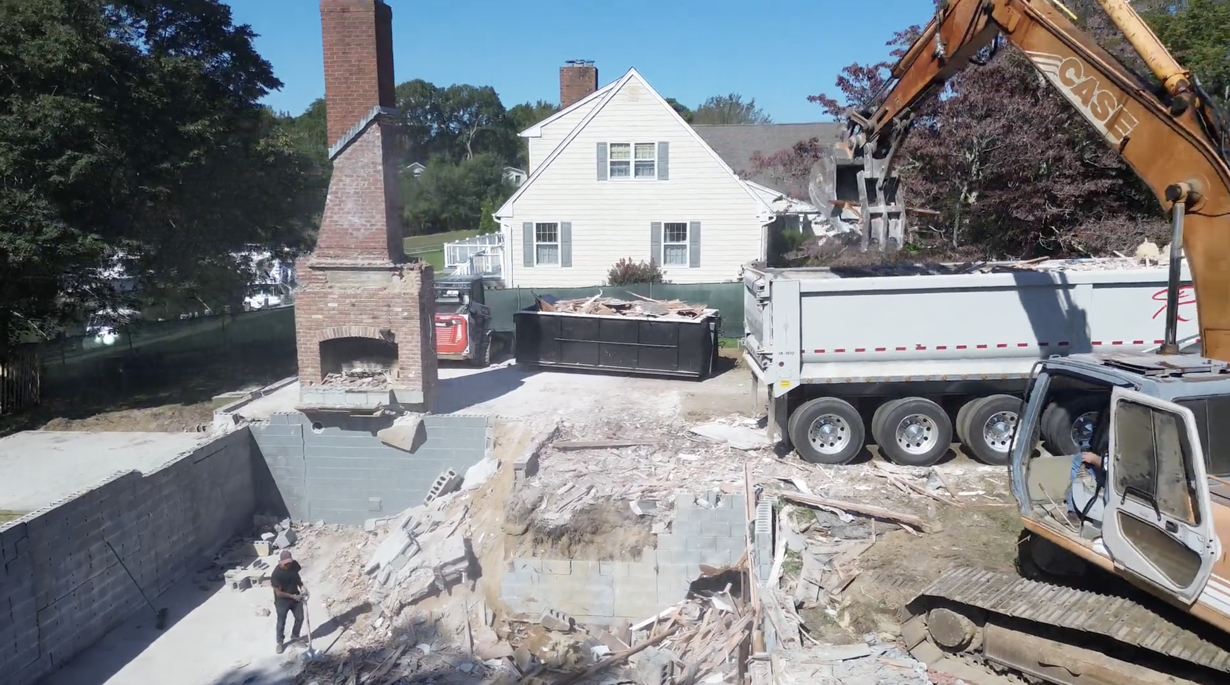 Construction site with bulldozer and truck demolishing a brick chimney and wall near a white house with gray shutters.