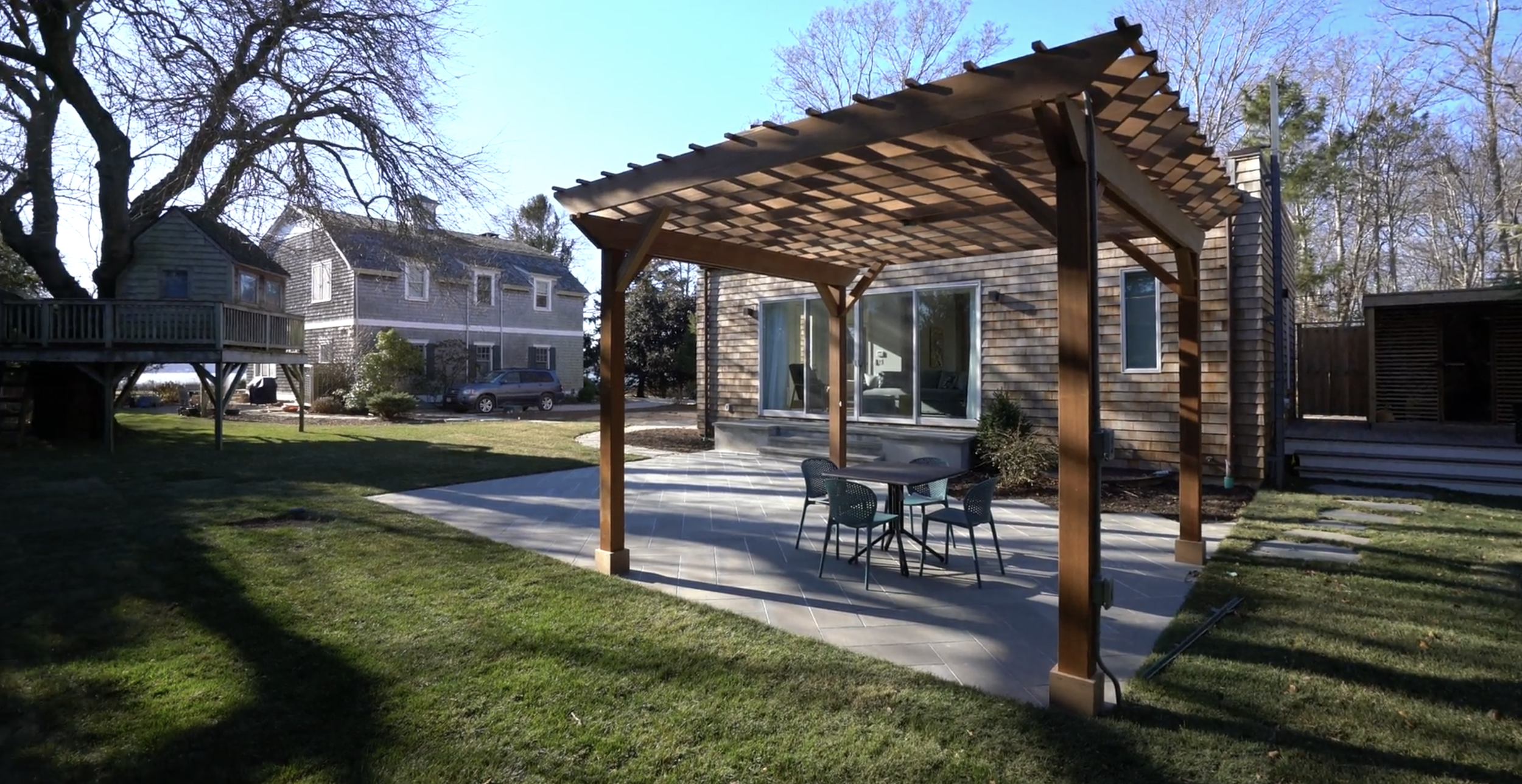 Backyard patio with a wooden pergola, outdoor dining table with four chairs, and sliding glass doors leading into a house. There is a grassy lawn, trees, and neighboring houses in the background.
