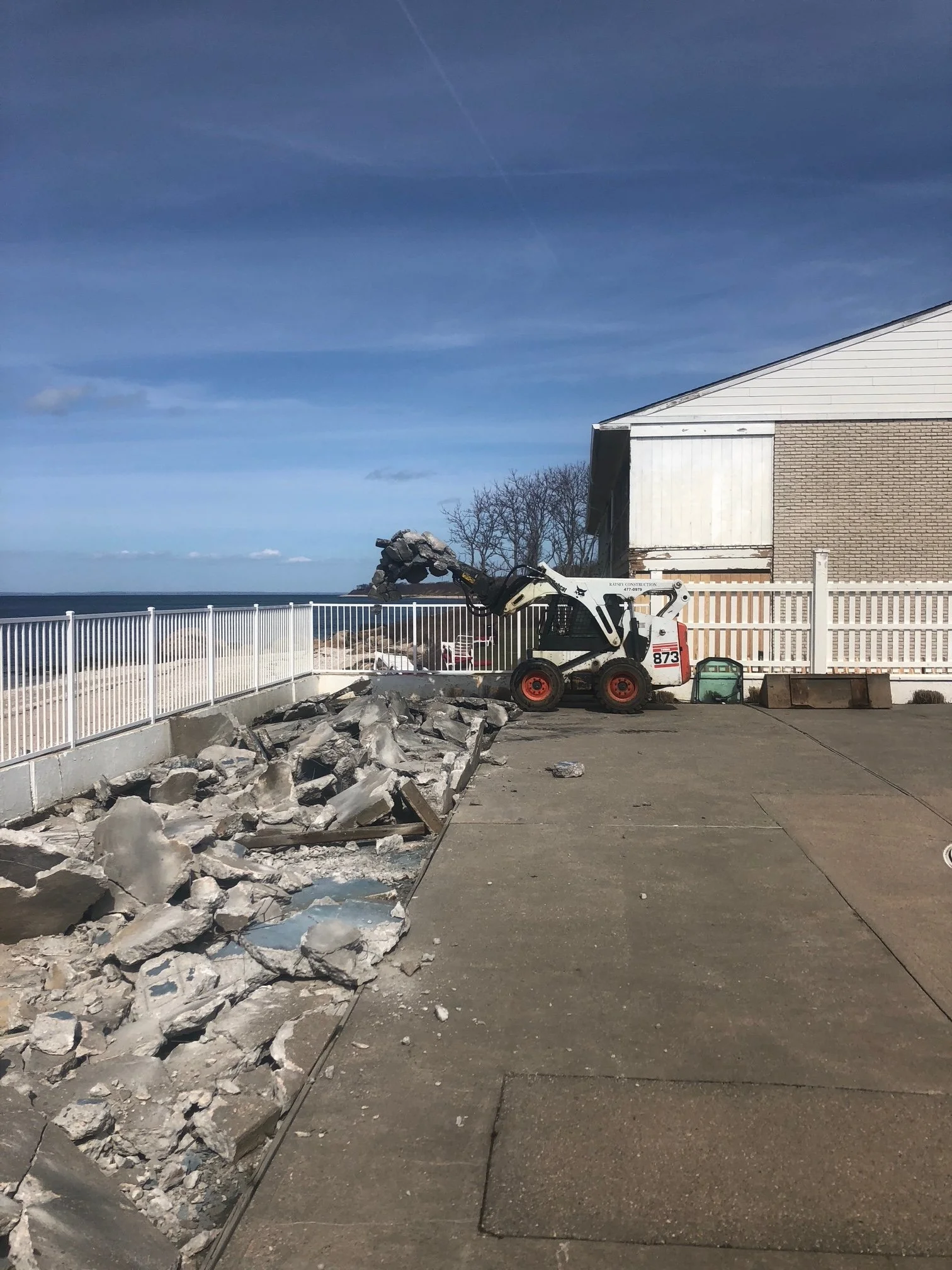 Construction equipment demolishing a concrete wall near a building with a fence, overlooking the ocean, under a blue sky.