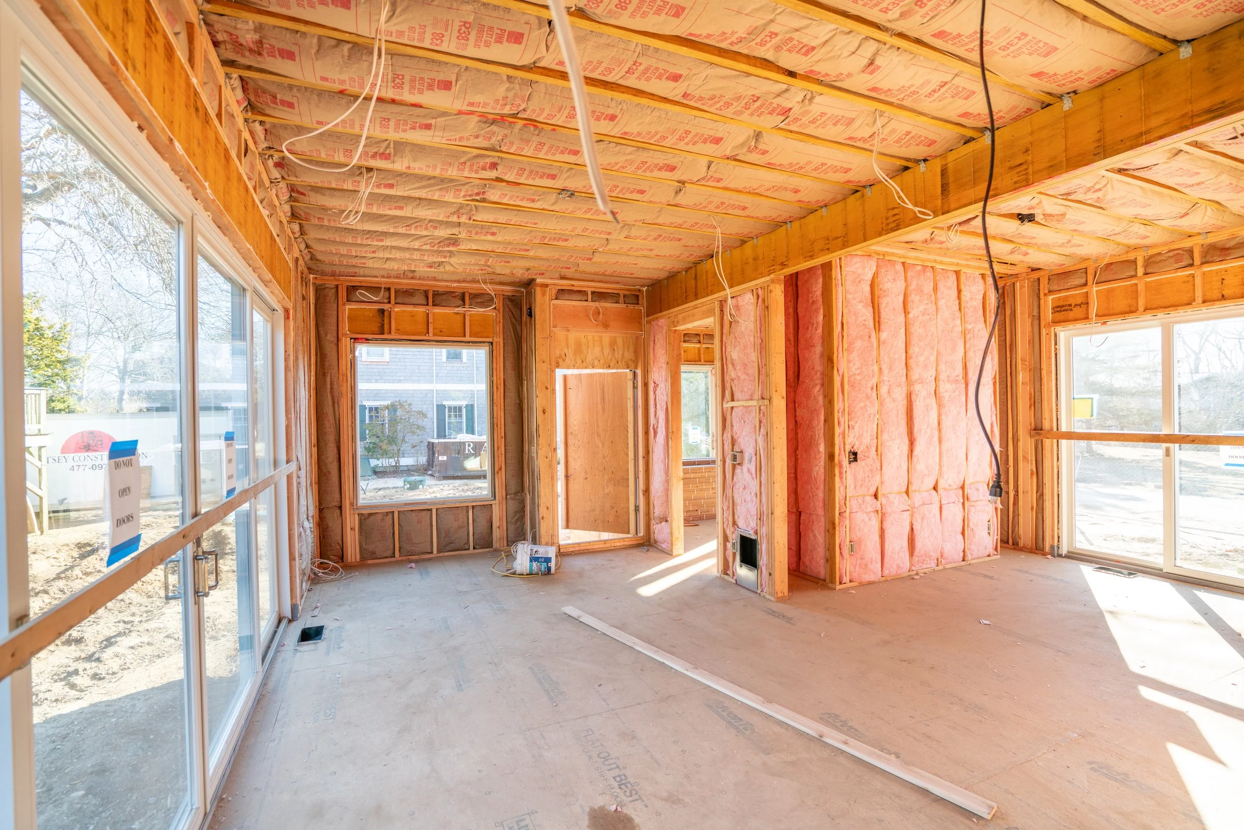 Interior view of a house under construction with exposed insulation, wooden framing, electrical wiring, and large windows.