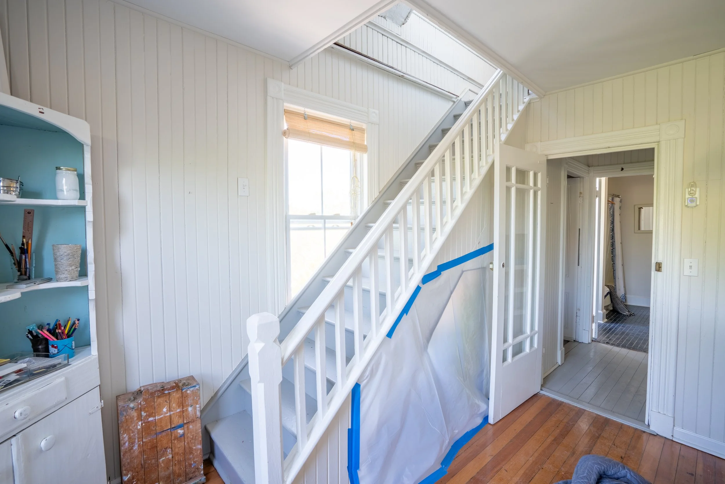 Interior of a house with a staircase, partially taped wall, white beadboard walls, a window, and a doorway leading to another room.