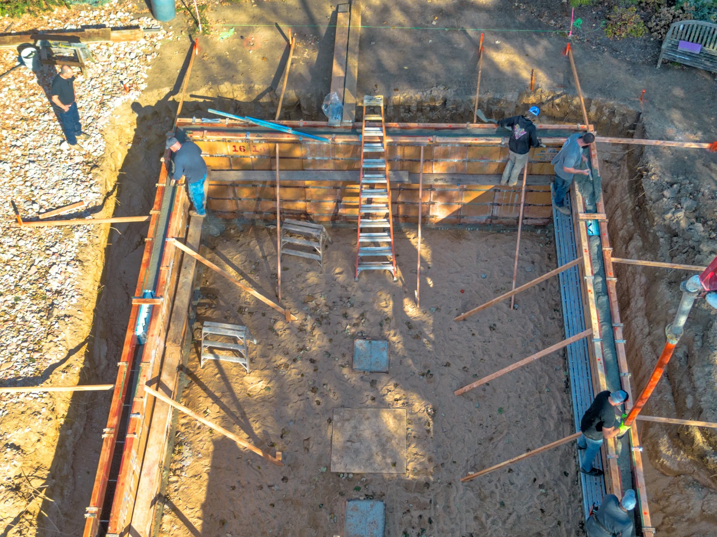 Construction workers working on a foundation in a rectangular excavation site with wooden supports and metal braces, with some tools and ladders inside.