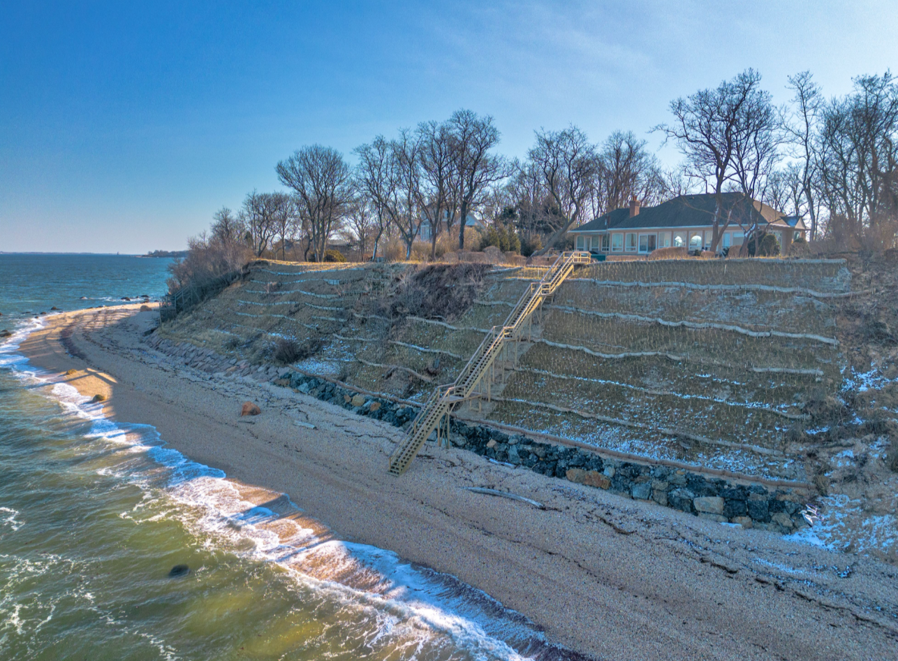 A house situated on a cliff overlooking a beach with stairs leading from the beach to the house, surrounded by leafless trees and a partly cloudy sky.