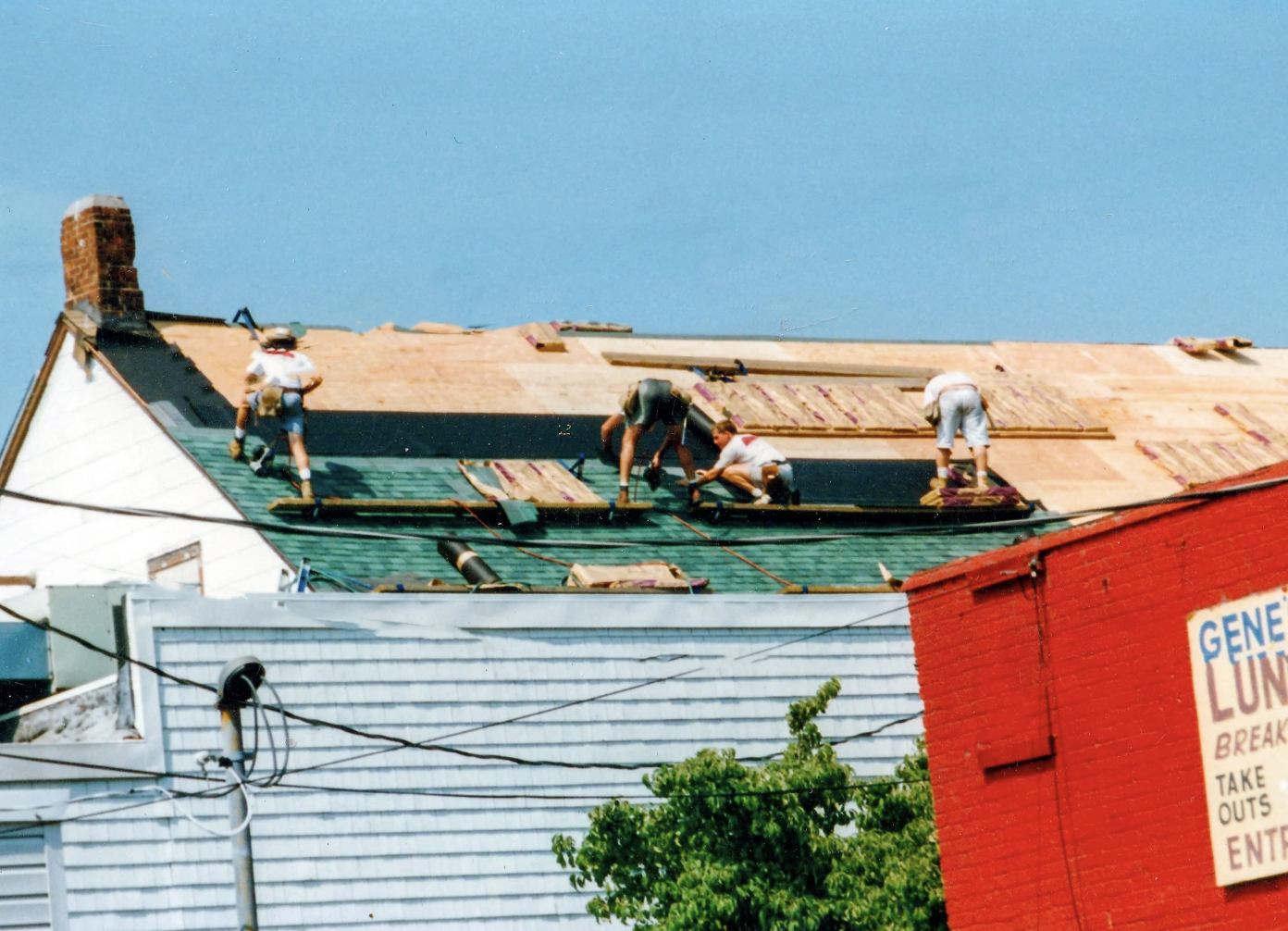 Ratsey Construction employees working on a house roof, replacing shingles, under a blue sky, with a red building on the right side.