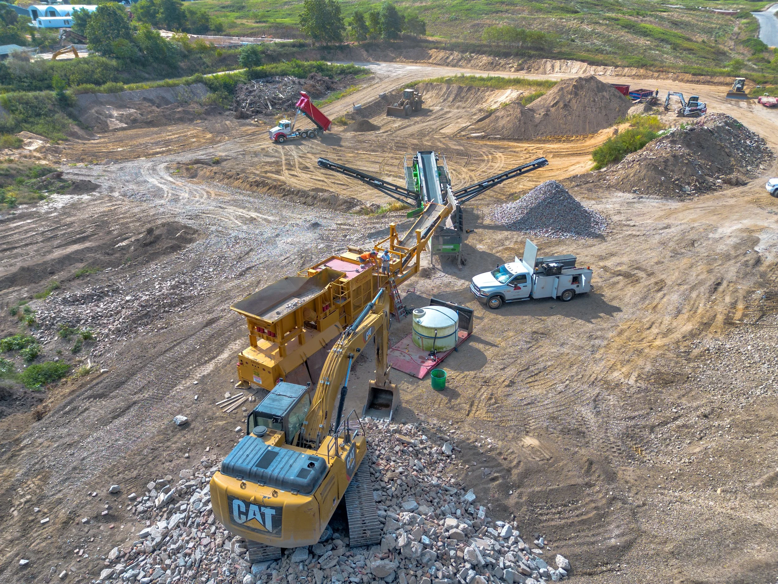 View of a construction site with a large yellow excavator moving rocks, a mobile crane, and pickup trucks, with dirt piles and machinery scattered around.
