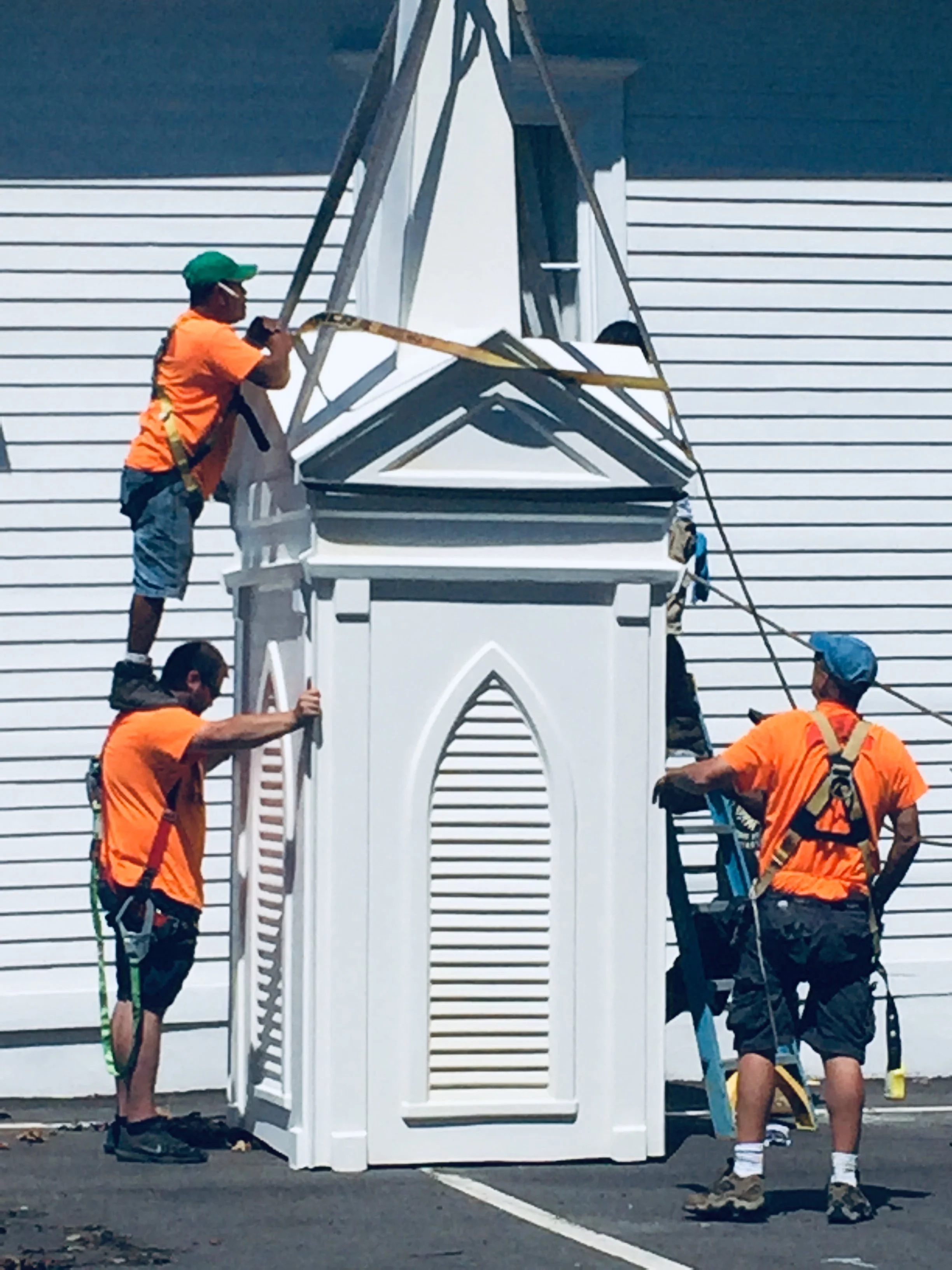 Four Ratsey workers installing a white church steeple on a building, wearing orange shirts and safety harnesses.