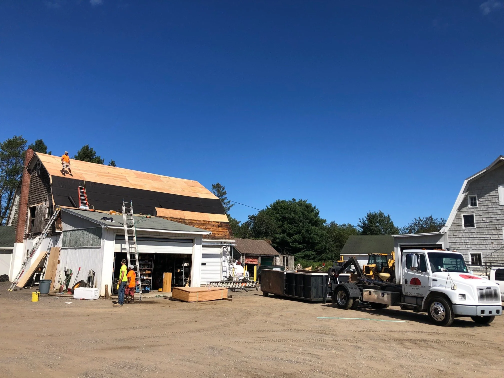 Construction workers installing a new roof on a house, with a truck parked nearby and ladders leaning against the house. The sky is clear and blue.