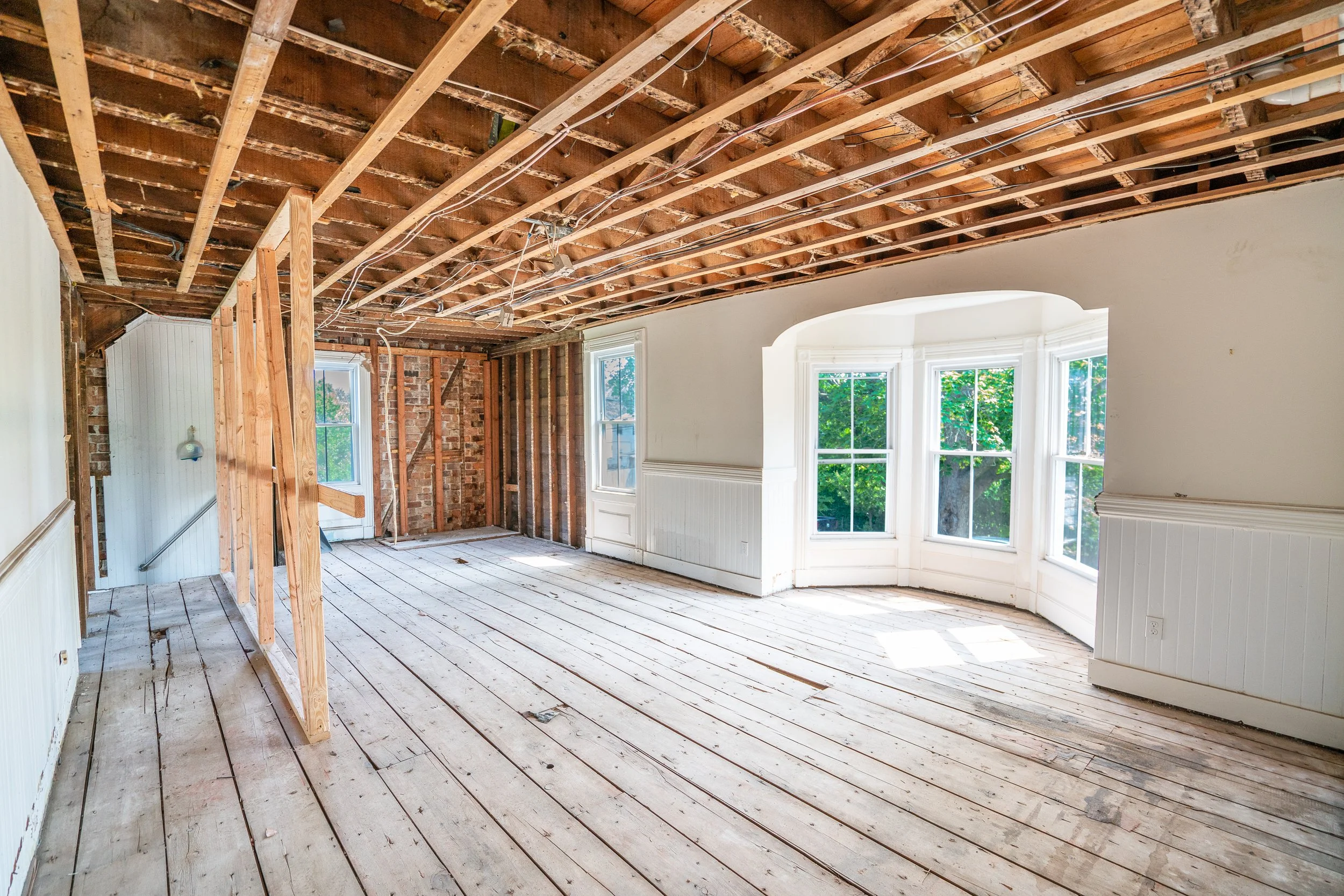 Room undergoing renovation with exposed ceiling beams and wood floor. A bay window with white trim and a view of green trees outside.