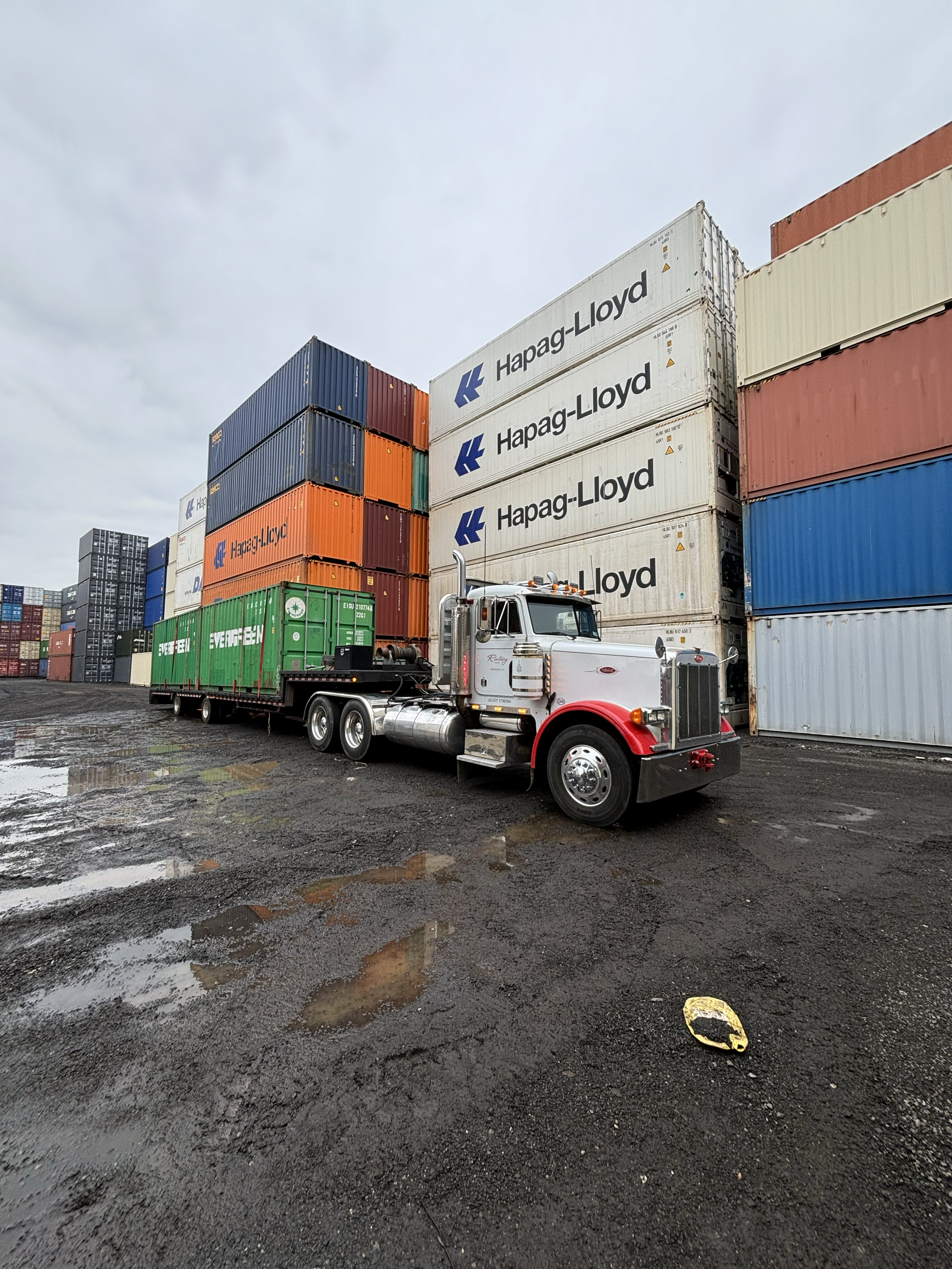 A semi-truck with a white cab and red accents parked on a muddy surface next to stacked shipping containers at a port or industrial area.