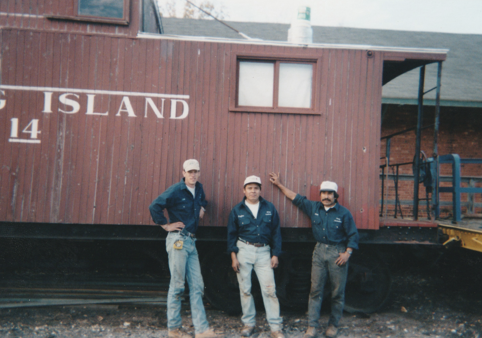 Three Ratsey workers in blue uniforms and white caps standing in front of a large red wooden building with the words 'ISLAND' visible, one worker with his hand on the building, on train tracks.