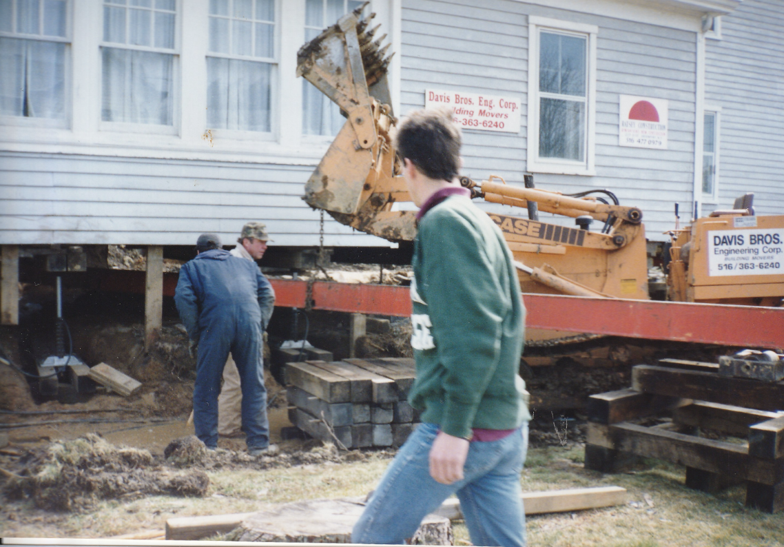 Construction workers and a young man at a house foundation site with a backhoe loader lifting soil or debris, in front of a gray house with multiple windows.