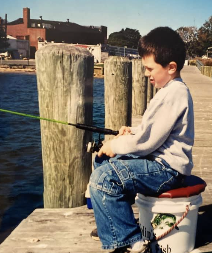 A young boy fishing off a wooden dock by the water, sitting on a white bucket with a red lid, with houses and trees in the background on a sunny day.