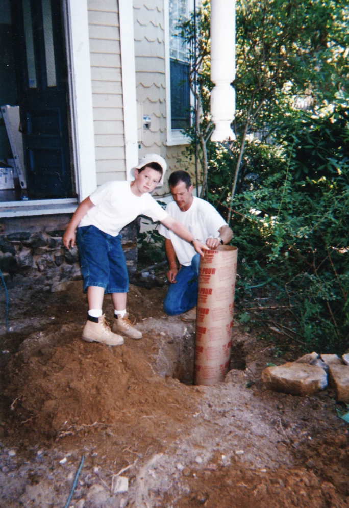 A boy and a man working near a house, installing a large pipe into the ground, with trees and a house exterior in the background.