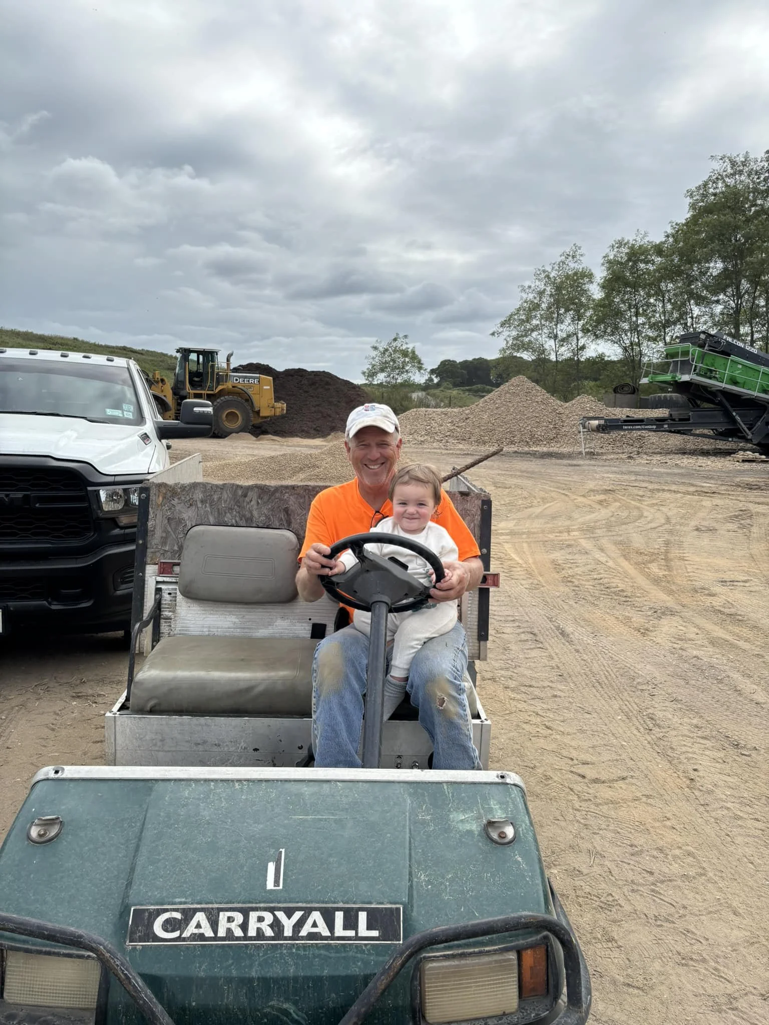 A man and a young girl smiling while sitting on a golf cart at a construction site with heavy machinery and piles of dirt in the background.