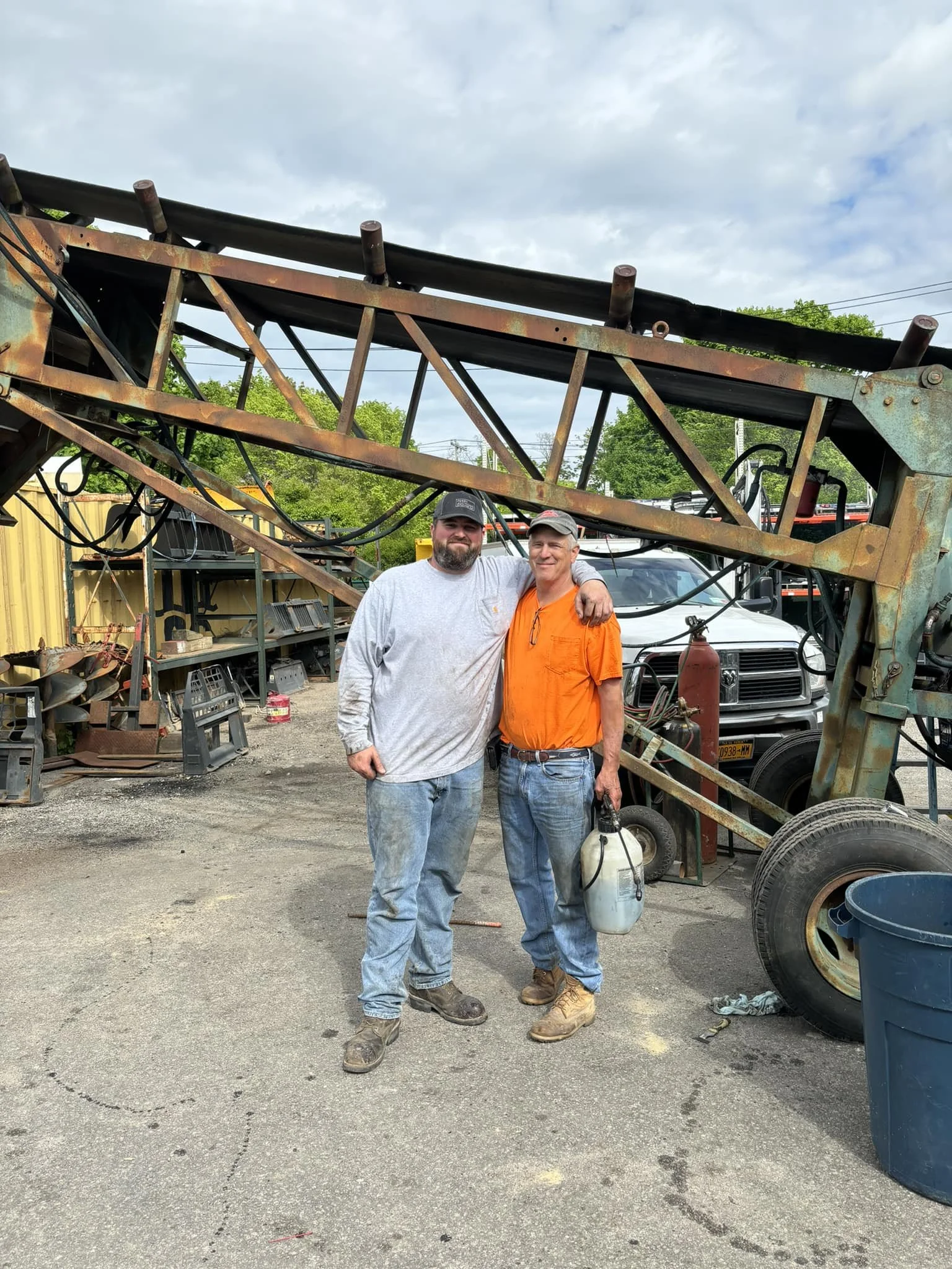 Two men standing together in front of large machinery outdoors. One man is in a gray long-sleeve shirt and jeans, the other in an orange shirt and jeans, holding a spray tank. They are smiling and have their arms around each other.