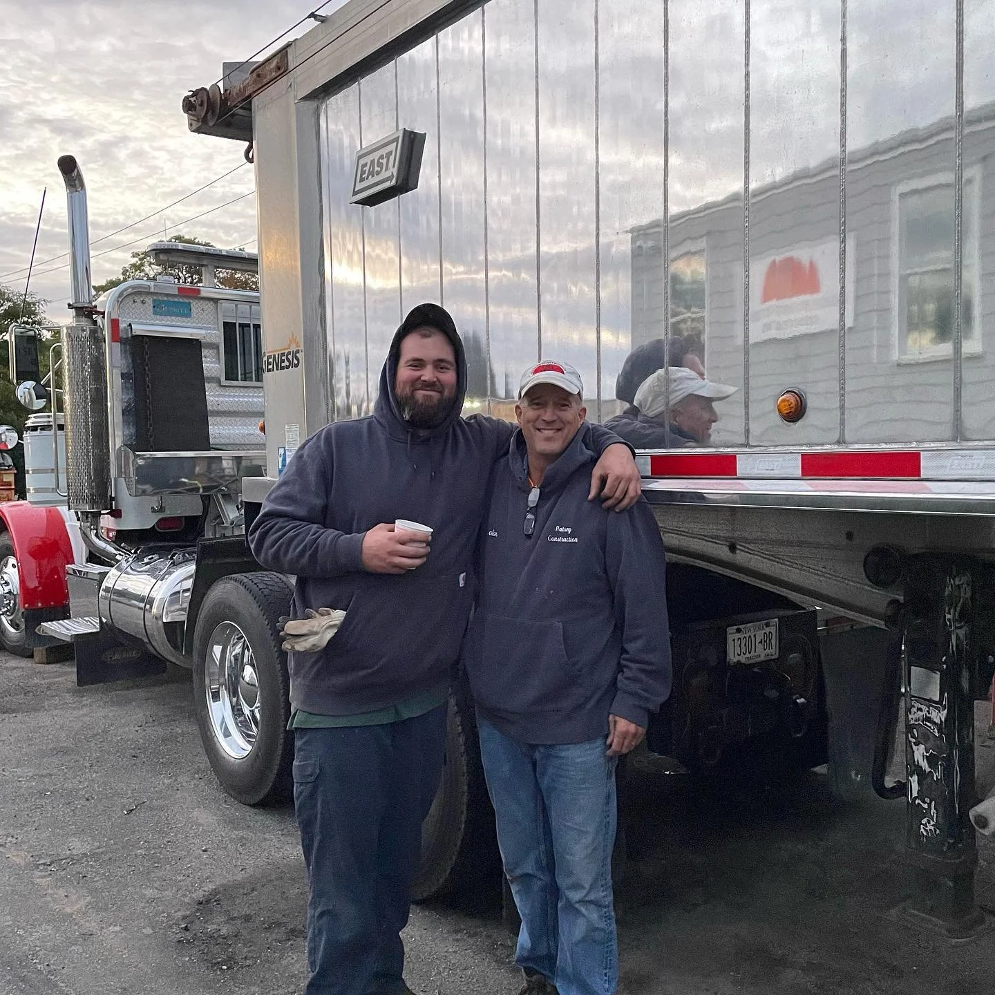 Colin Ratsey Senior  and Colin Ratsey Jr smiling and standing in front of a large truck, with one man holding a cup. They are outdoors, wearing hoodies, and appear to be happy.