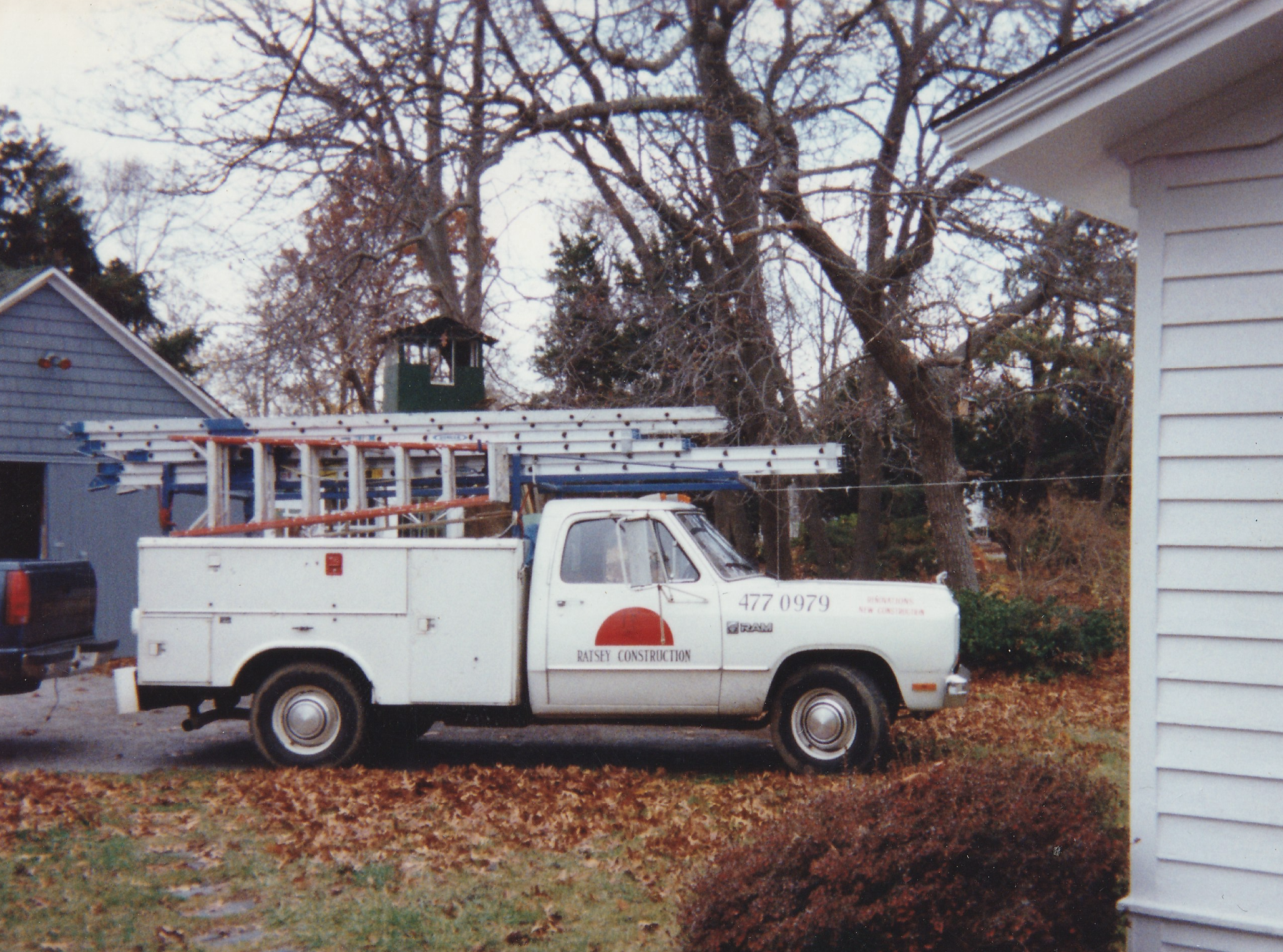 A white utility truck with ladders on top, parked outdoors on a residential yard with fallen leaves. The truck has a red and white logo for 'Rasley Construction' and phone number '477 0979' on the door. Surrounding trees are mostly bare.