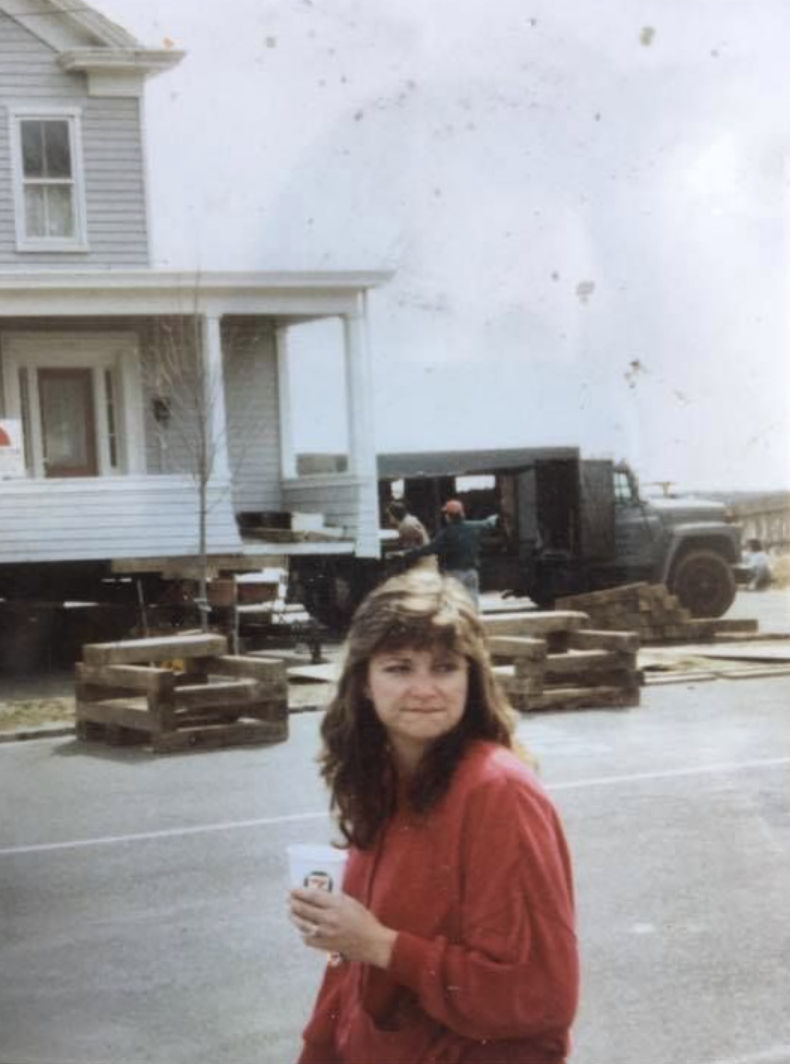 A woman with wavy brown hair wearing a red jacket holding a paper cup, standing in front of a house under construction with wooden pallets and construction workers and vehicles in the background.