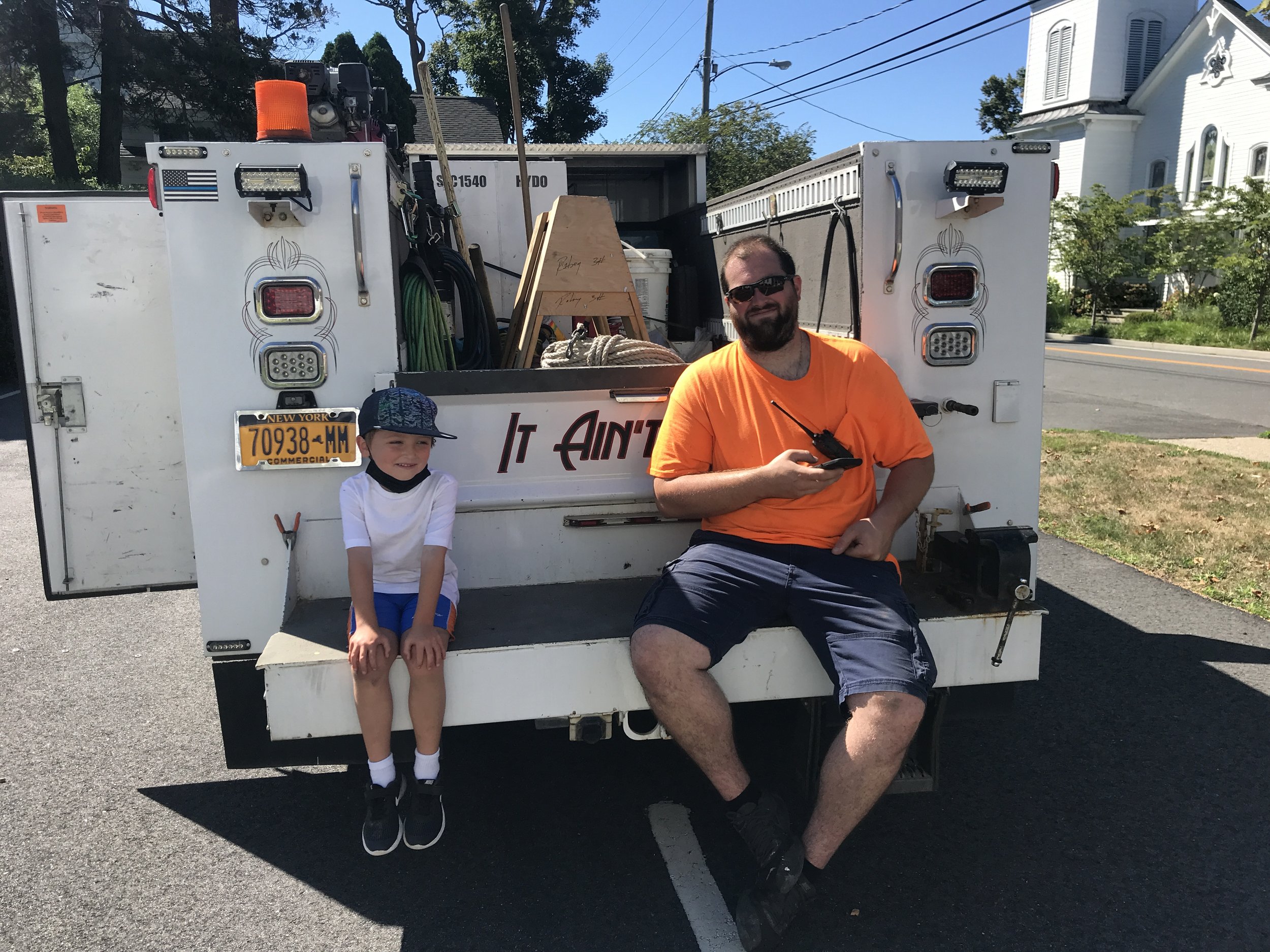 A man and a young boy sitting on the back of a utility truck parked on a street in a neighborhood. The man is wearing a bright orange T-shirt, dark shorts, and sunglasses, holding a walkie-talkie. The boy is wearing a white and blue outfit with a cap