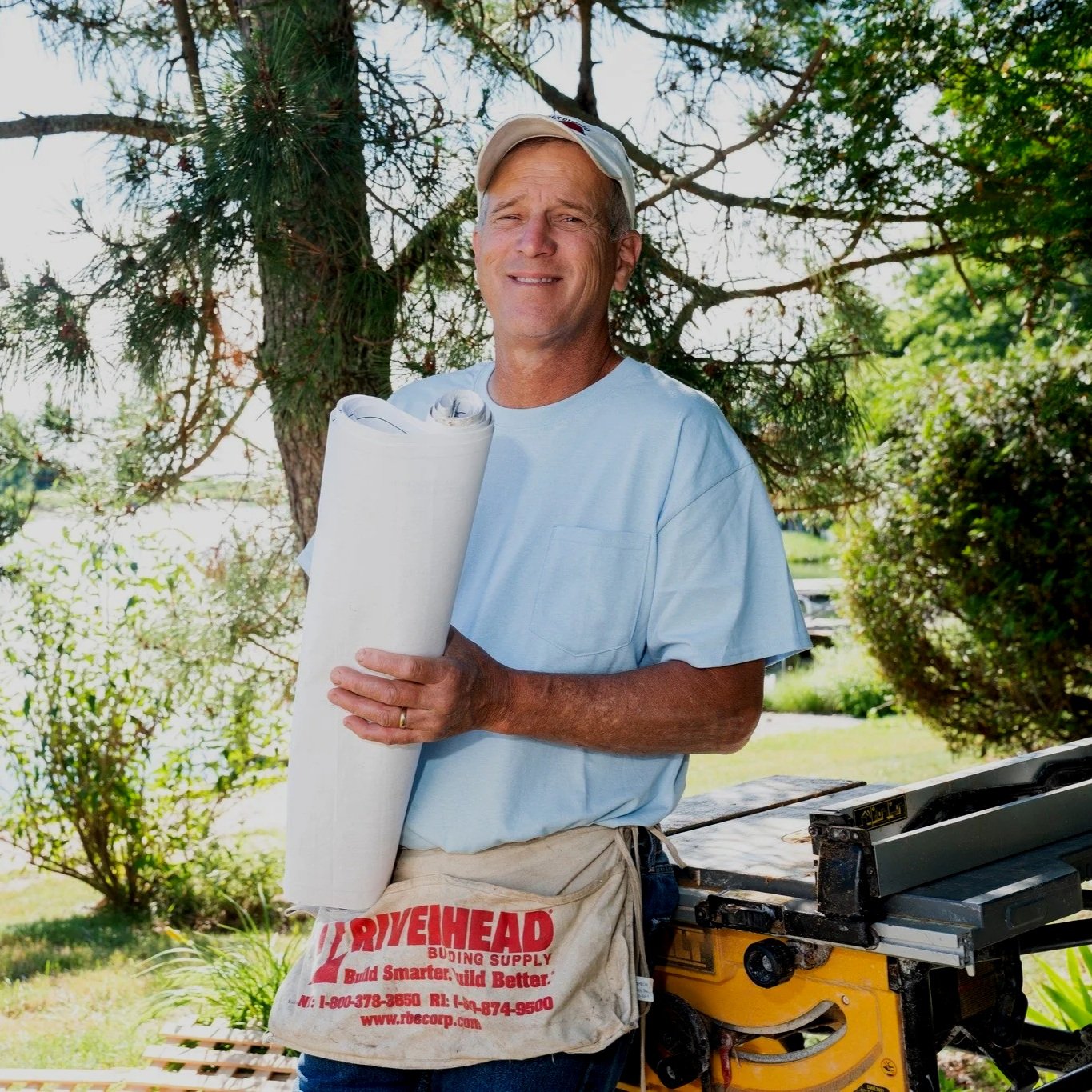 A man standing outdoors, holding a rolled-up blueprint in his arms, with a table saw next to him, surrounded by trees and greenery.