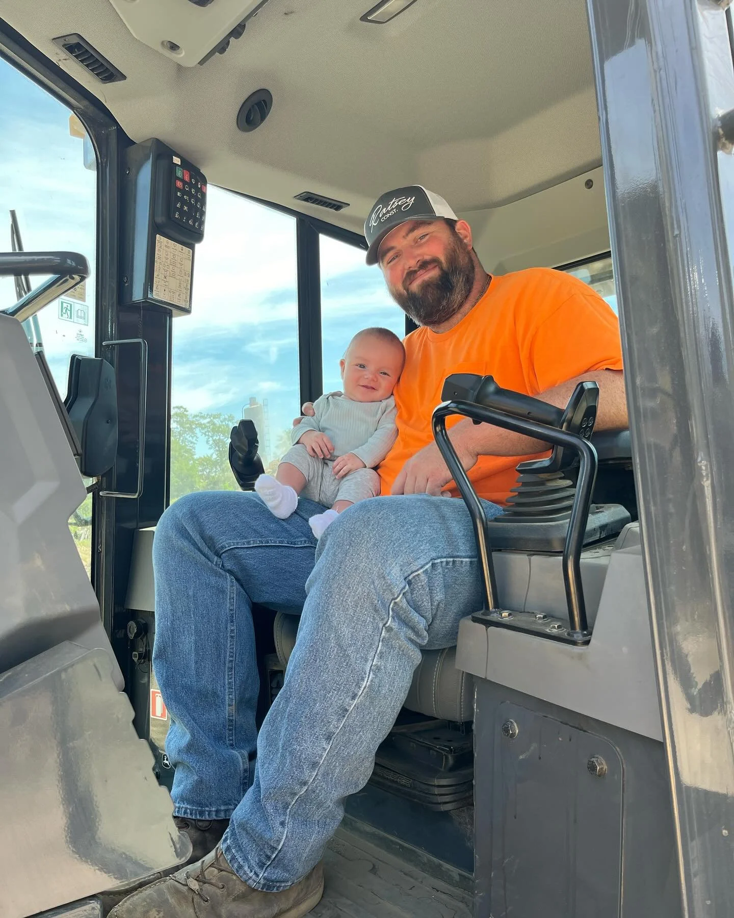 Man with a beard in an orange shirt and gray cap sitting in the cabin of a tractor, holding a smiling baby dressed in a light gray outfit, with a sunny sky and green trees visible outside the windows.
