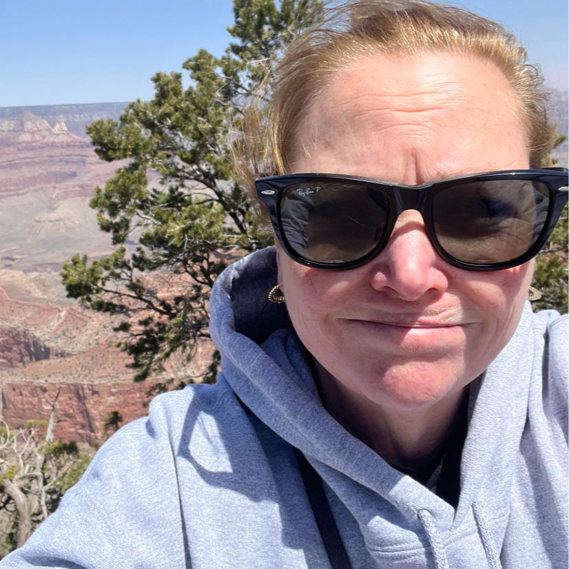 A woman with blonde hair wearing sunglasses and a gray hoodie taking a selfie at the Grand Canyon with trees and canyon cliffs in the background.