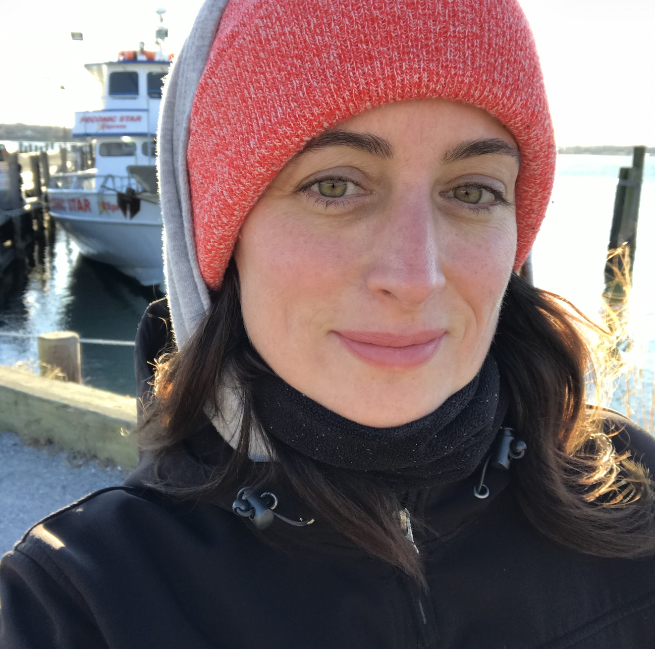 A woman taking a selfie at a marina, wearing a red beanie, black jacket, and black scarf, with docked boats in the background.