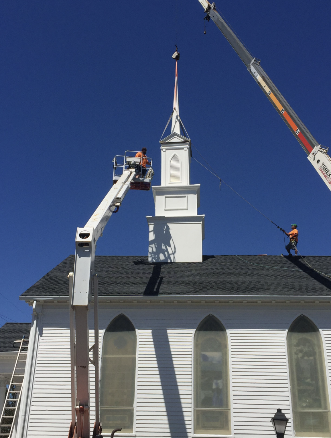 Workers in orange shirts and green helmets using cranes and a lift to work on the steeple of a white church with arched windows against a clear blue sky.