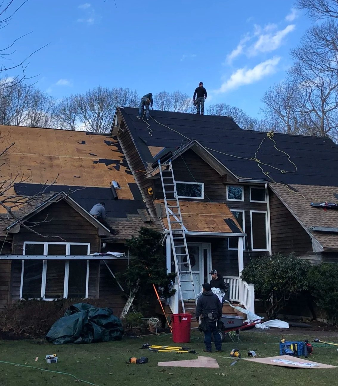 Workers installing new roof shingles on a house, with some in the process of removing old shingles and others laying new ones. Tools and equipment are scattered on the lawn.