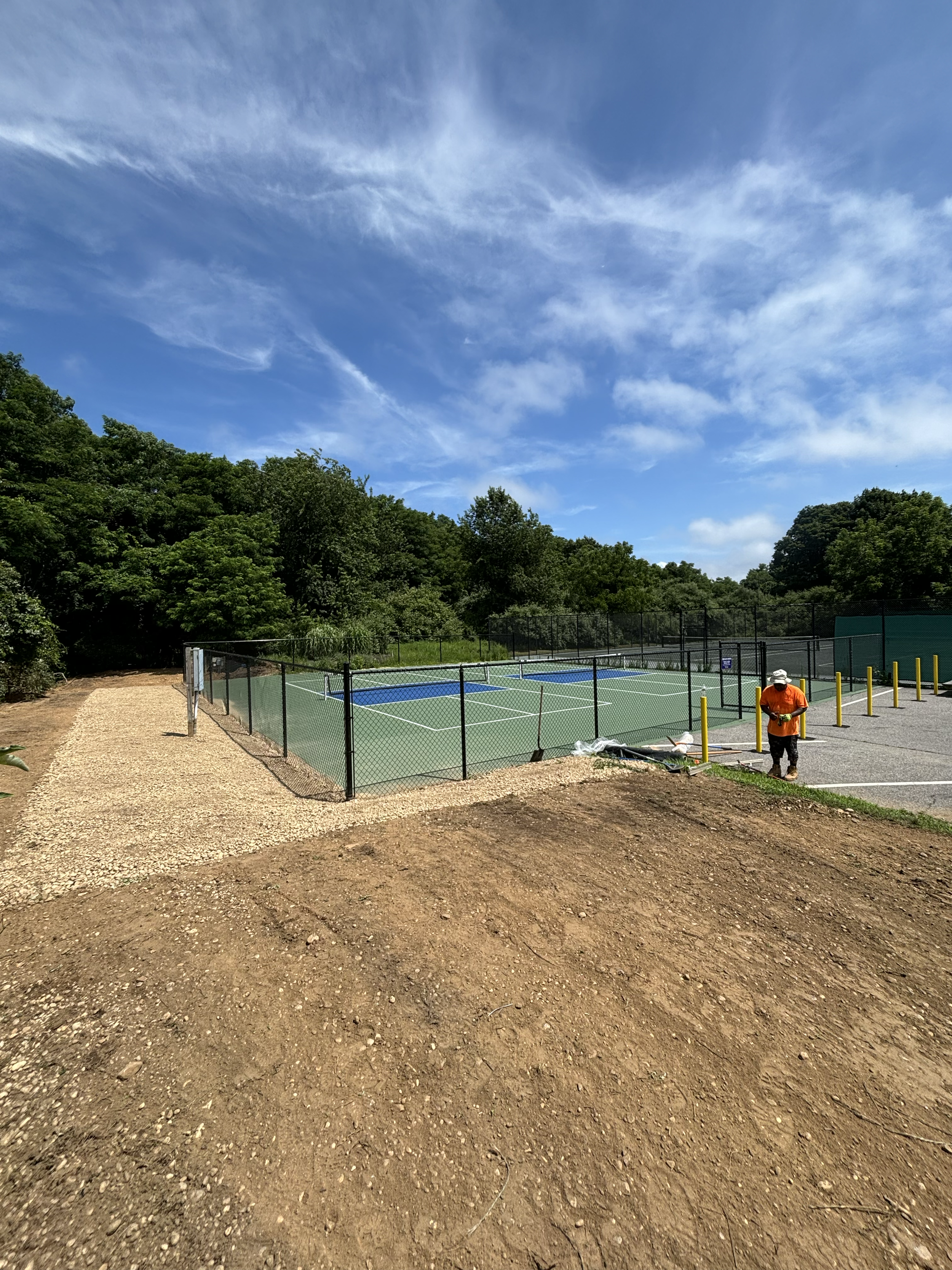 A tennis court under construction with a worker standing beside the fence, surrounded by trees and a blue sky with clouds.