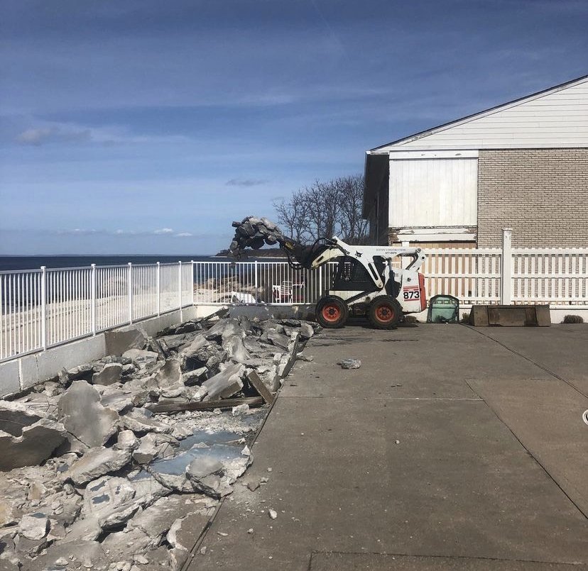 A small construction vehicle breaking up concrete on a paved outdoor area next to a white fence and a house, with rocks and debris scattered on the ground.