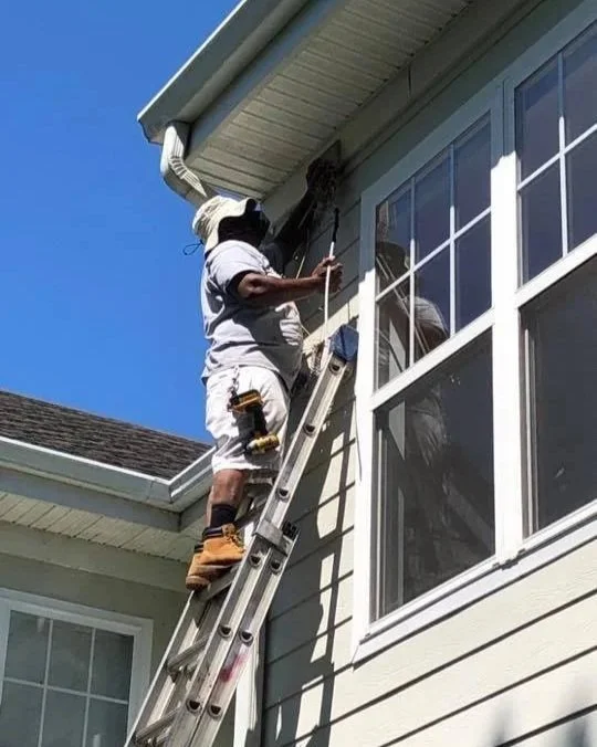 A Ratsey Construction worker on a ladder outside a house, fixing or inspecting the house gutter near a window under a clear blue sky.