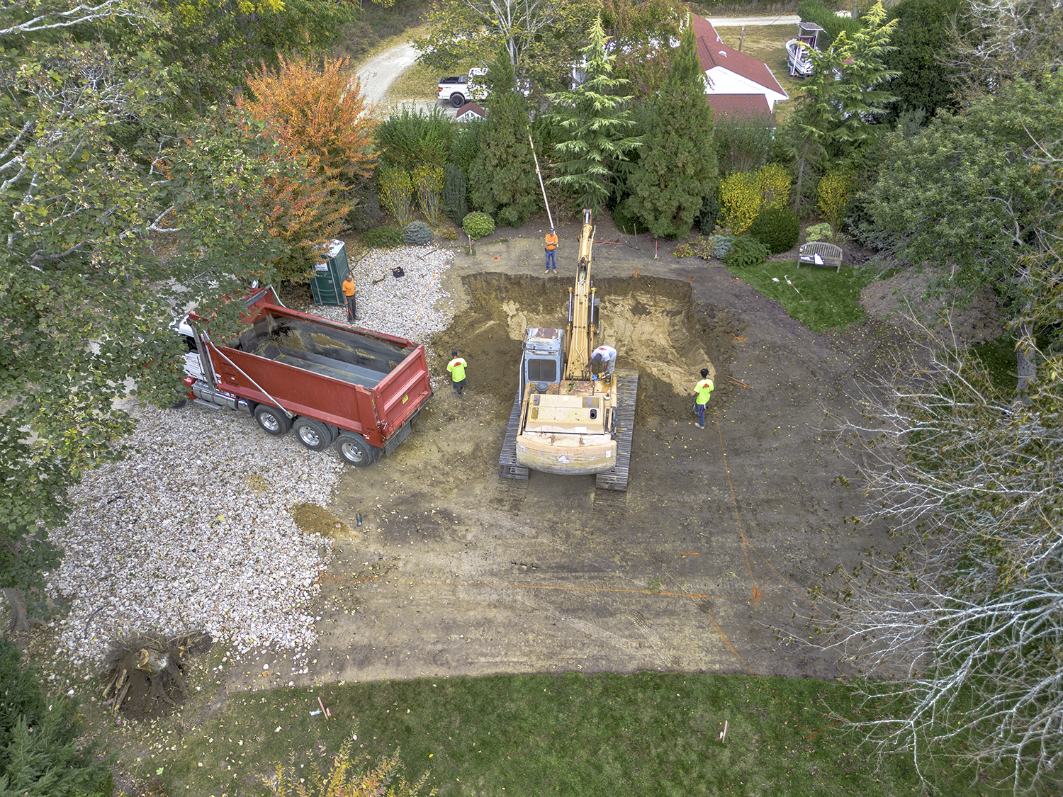 Ratsey construction site with a large excavator digging a hole, a red dump truck nearby, and several workers in bright clothing. The area is surrounded by trees and residential buildings.
