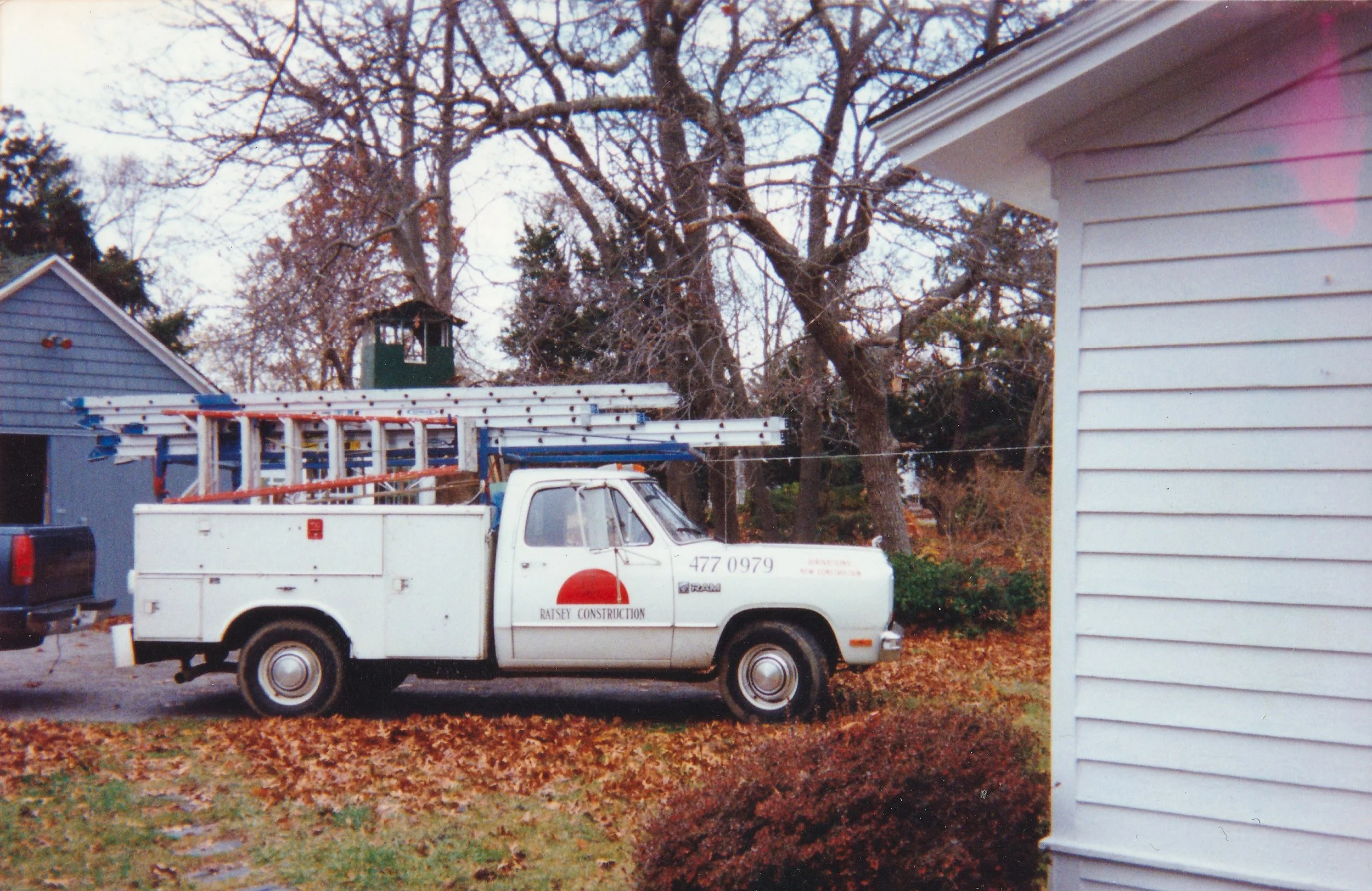A while utility truck with a red and black logo and the words 'Ratsey Construction' parked on a street surrounded by fall leaves. The truck has a ladder mounted on top. 