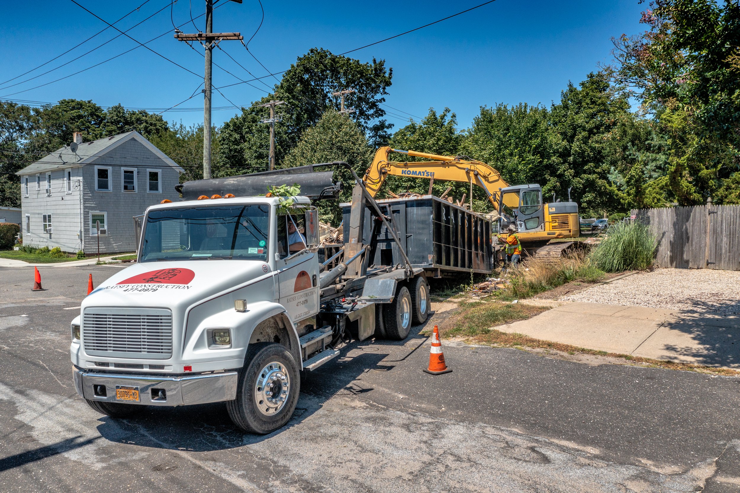 Ratsey construction site with a white construction truck and a yellow excavator working on a sidewalk, with workers in safety gear, orange cones, and a residential neighborhood in the background.