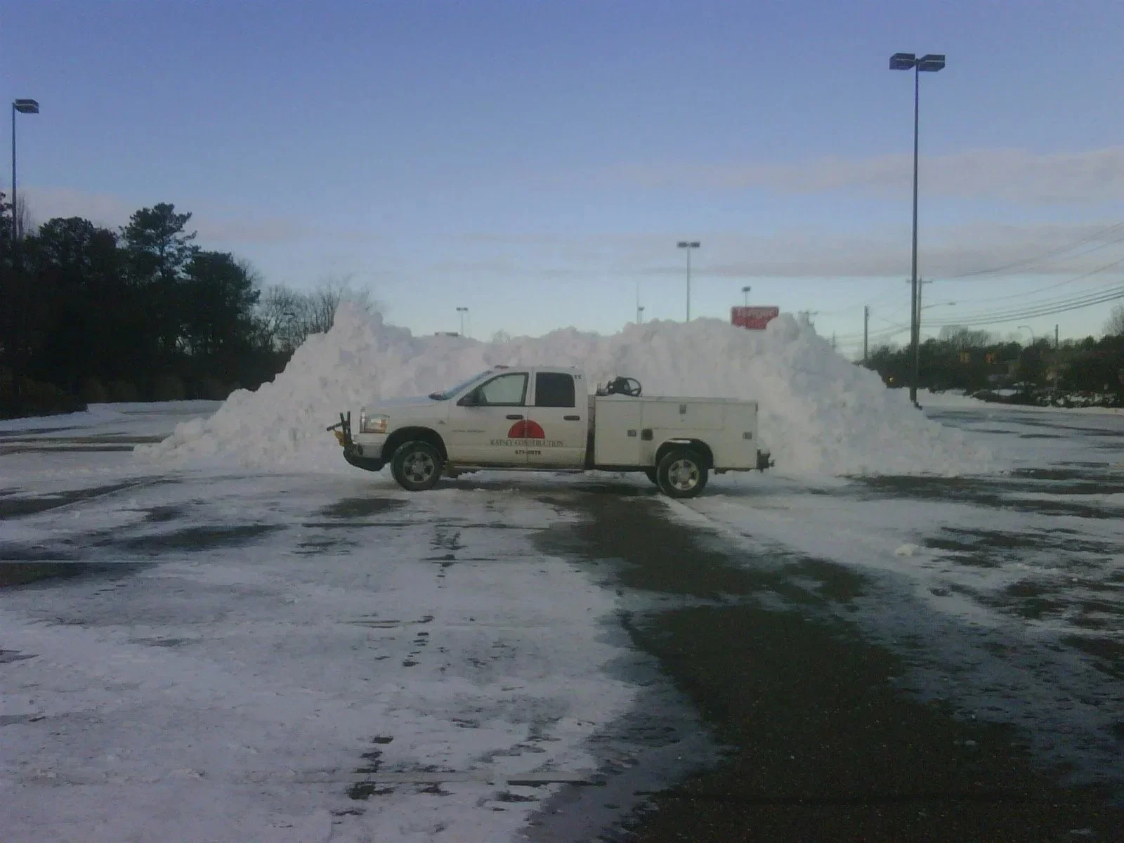 A parking lot covered in snow with a pickup truck from Ratsey Construction parked in front of a large snow pile, four tall light poles, trees in the background, and cloudy sky.