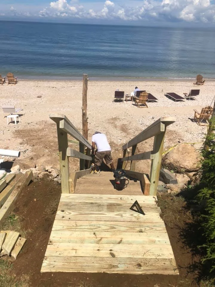 View from top of wooden staircase leading down to a sandy beach with lounge chairs and a few people near the shoreline, with calm ocean water and cloudy sky in the background.